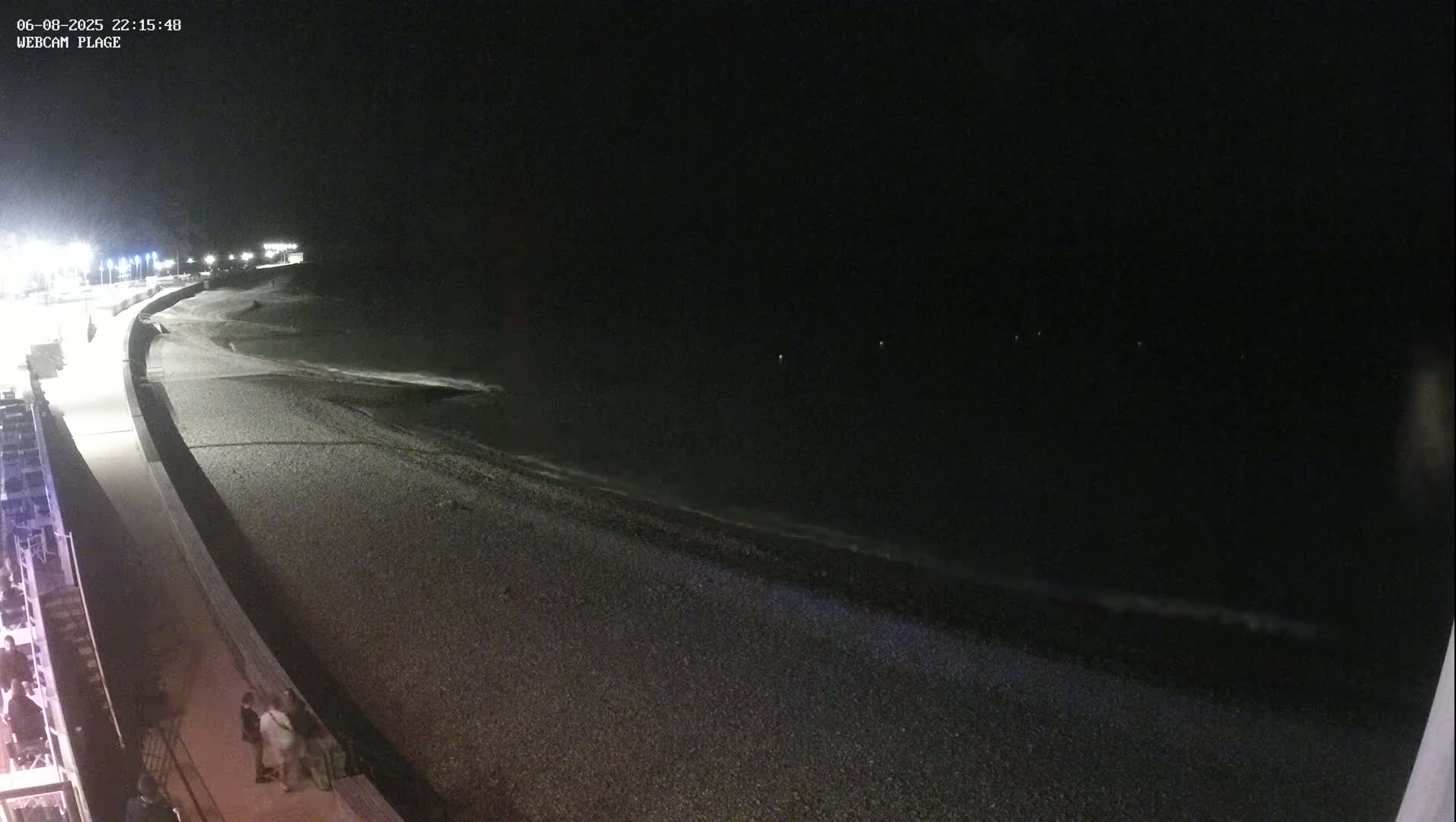 A dark, nighttime beach scene shows a pebbled shore with gentle waves lapping, a paved walkway lit by lamps, and a few people walking along the path.