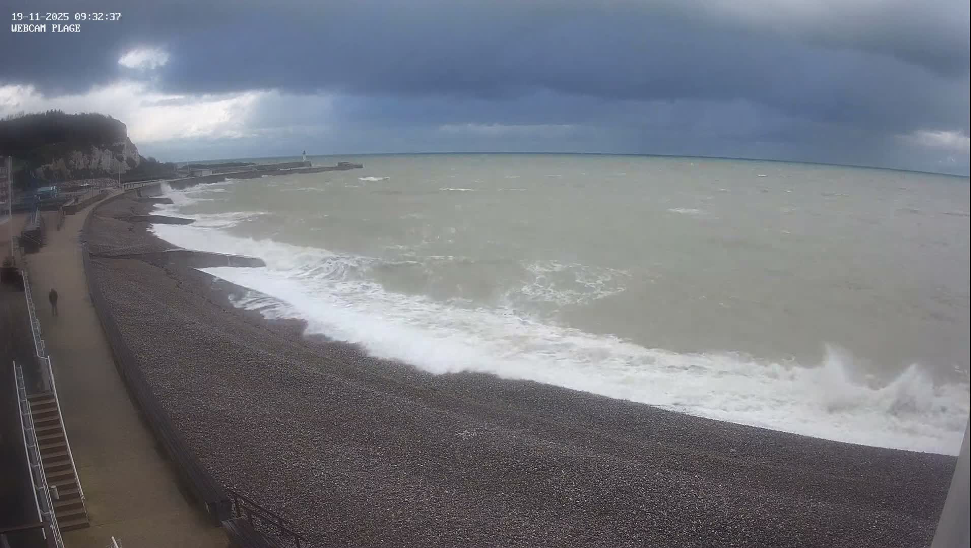 A rough, grey sea with white waves crashes onto a pebble beach under a dark, overcast sky, while a lone figure walks along a promenade next to distant cliffs and a lighthouse.