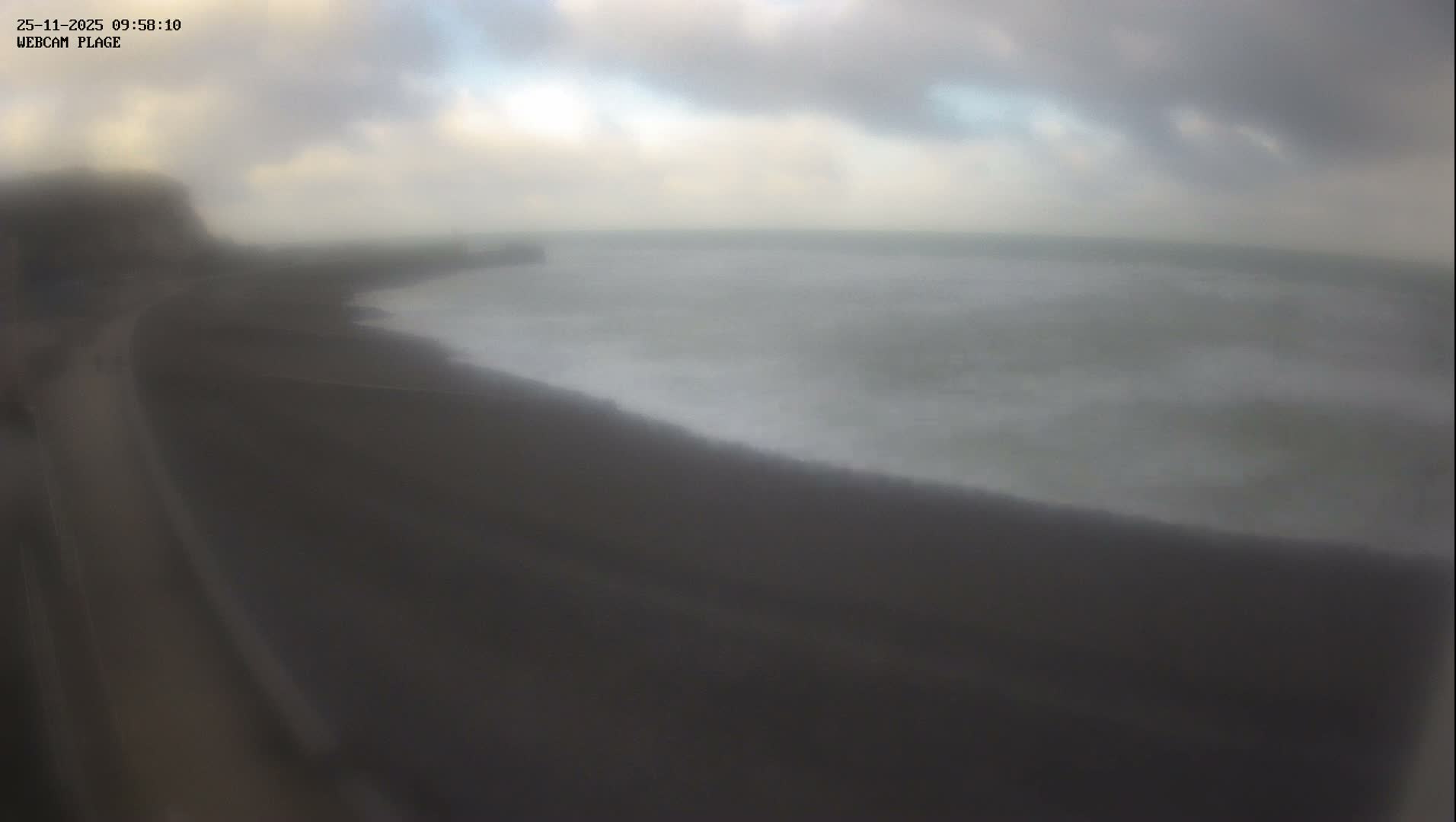A hazy and overcast day is depicted, showing a dark beach or paved area curving along a coastline with the ocean stretching to the horizon under a cloudy sky.