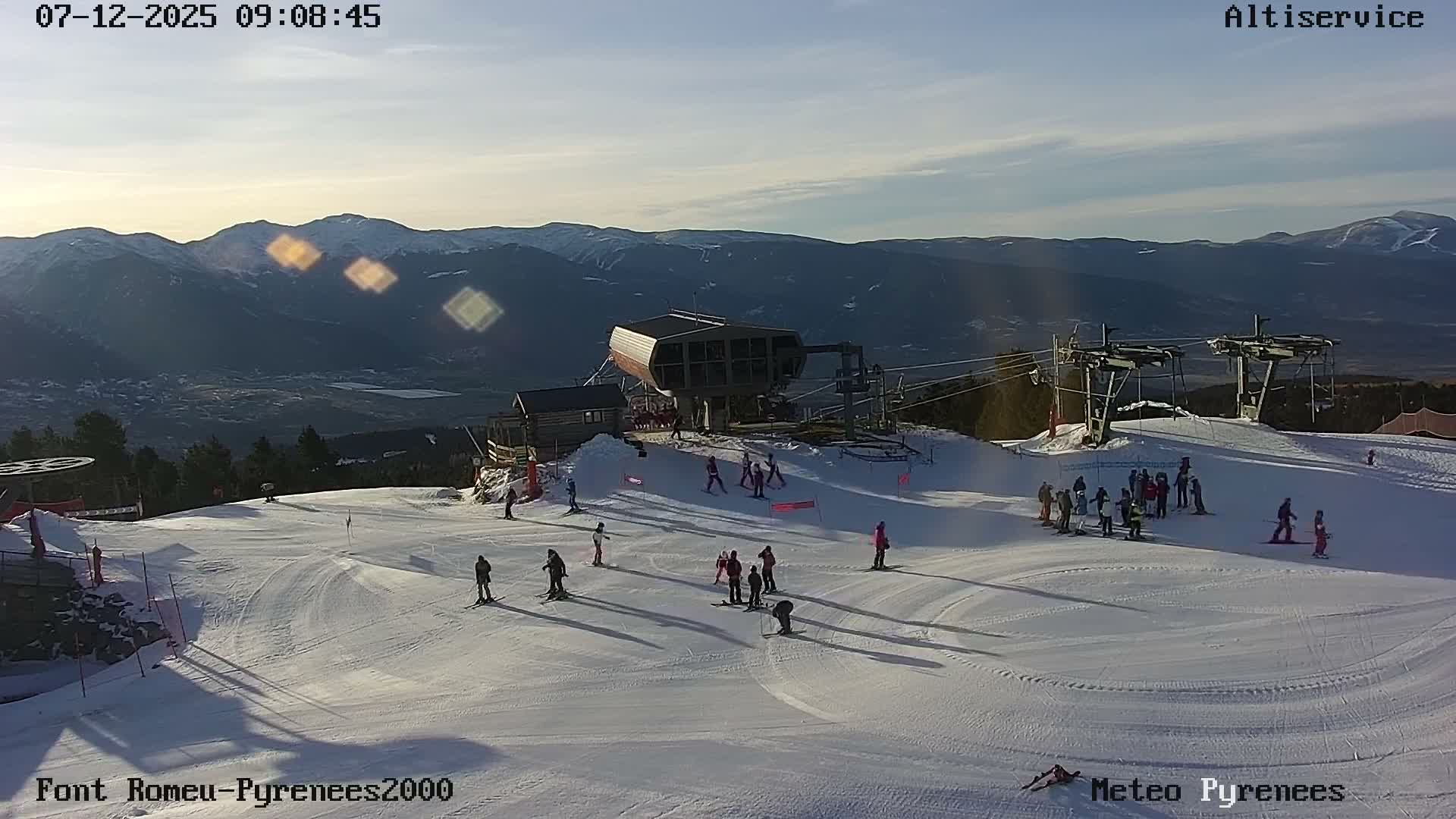Numerous skiers are visible on a sunlit, snowy mountain slope at a bustling ski resort, with ski lifts overhead and distant snow-capped mountains under a bright, partly cloudy sky on a clear winter day.