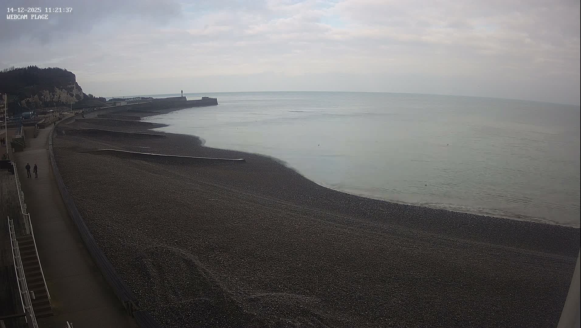 Numerous skiers are visible on a sunlit, snowy mountain slope at a bustling ski resort, with ski lifts overhead and distant snow-capped mountains under a bright, partly cloudy sky on a clear winter day.