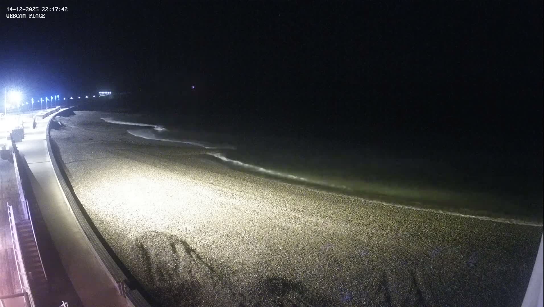 Numerous skiers are visible on a sunlit, snowy mountain slope at a bustling ski resort, with ski lifts overhead and distant snow-capped mountains under a bright, partly cloudy sky on a clear winter day.