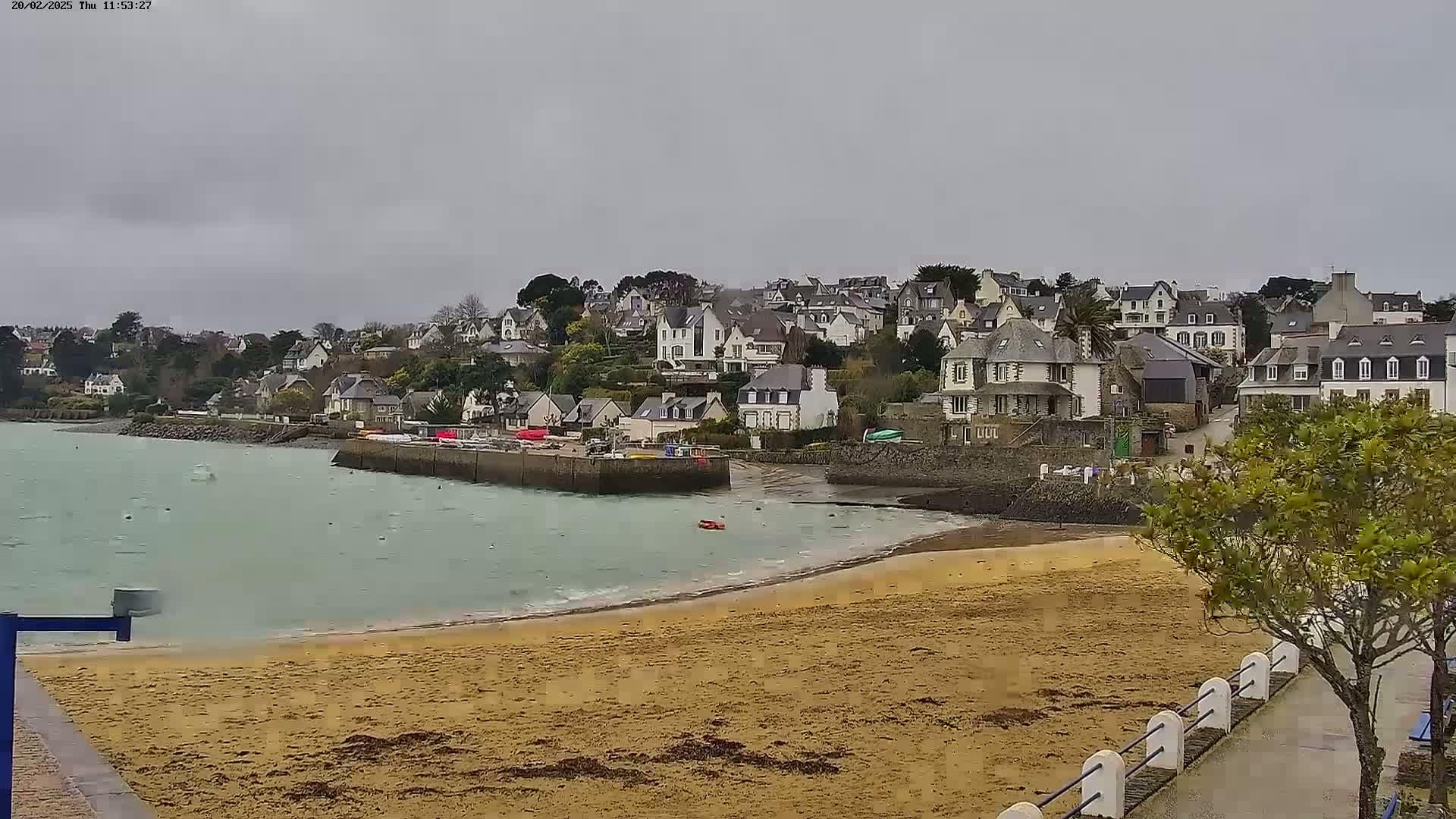 A sandy beach curves around a calm bay with a small harbor and a village of white buildings on a hill under an overcast sky.