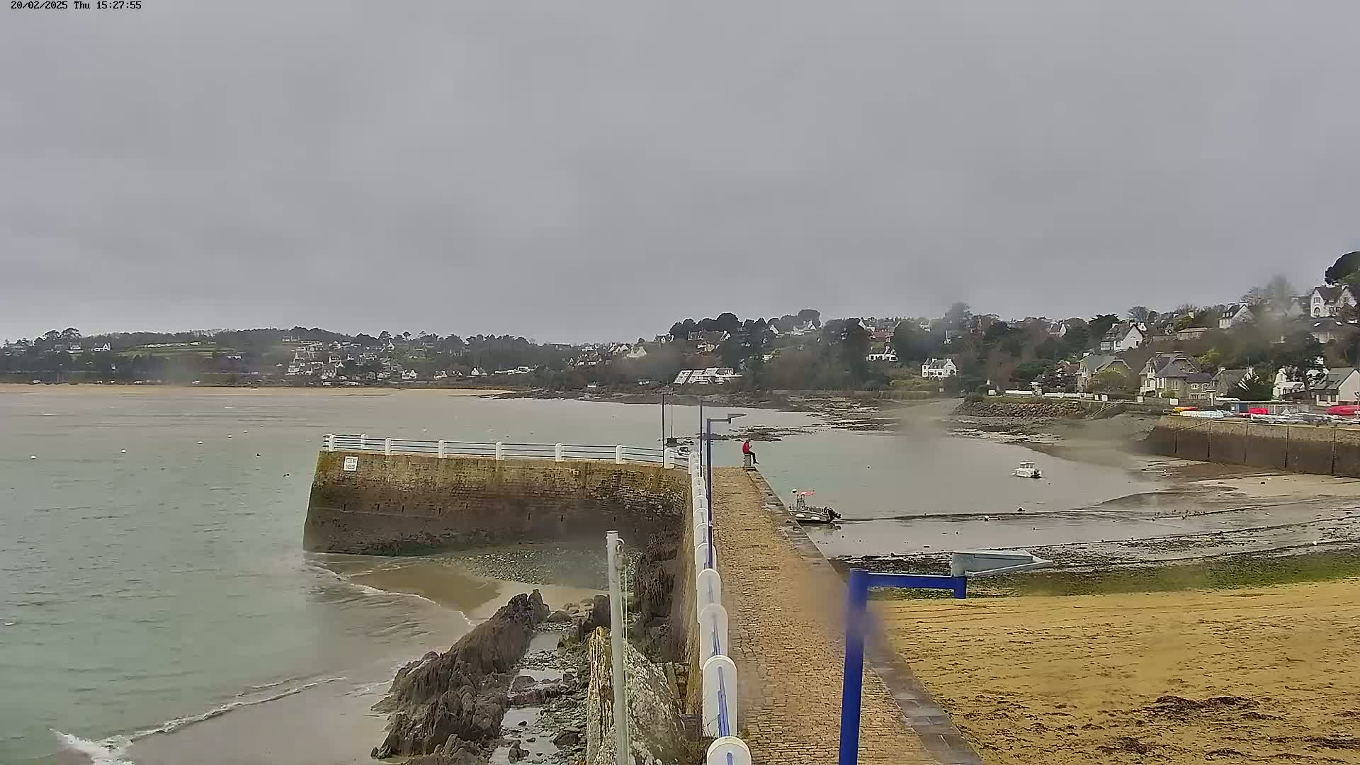 A person sits on a stone pier overlooking a calm, gray-toned bay, with a small boat in the shallows and a village nestled on the hillside beyond.