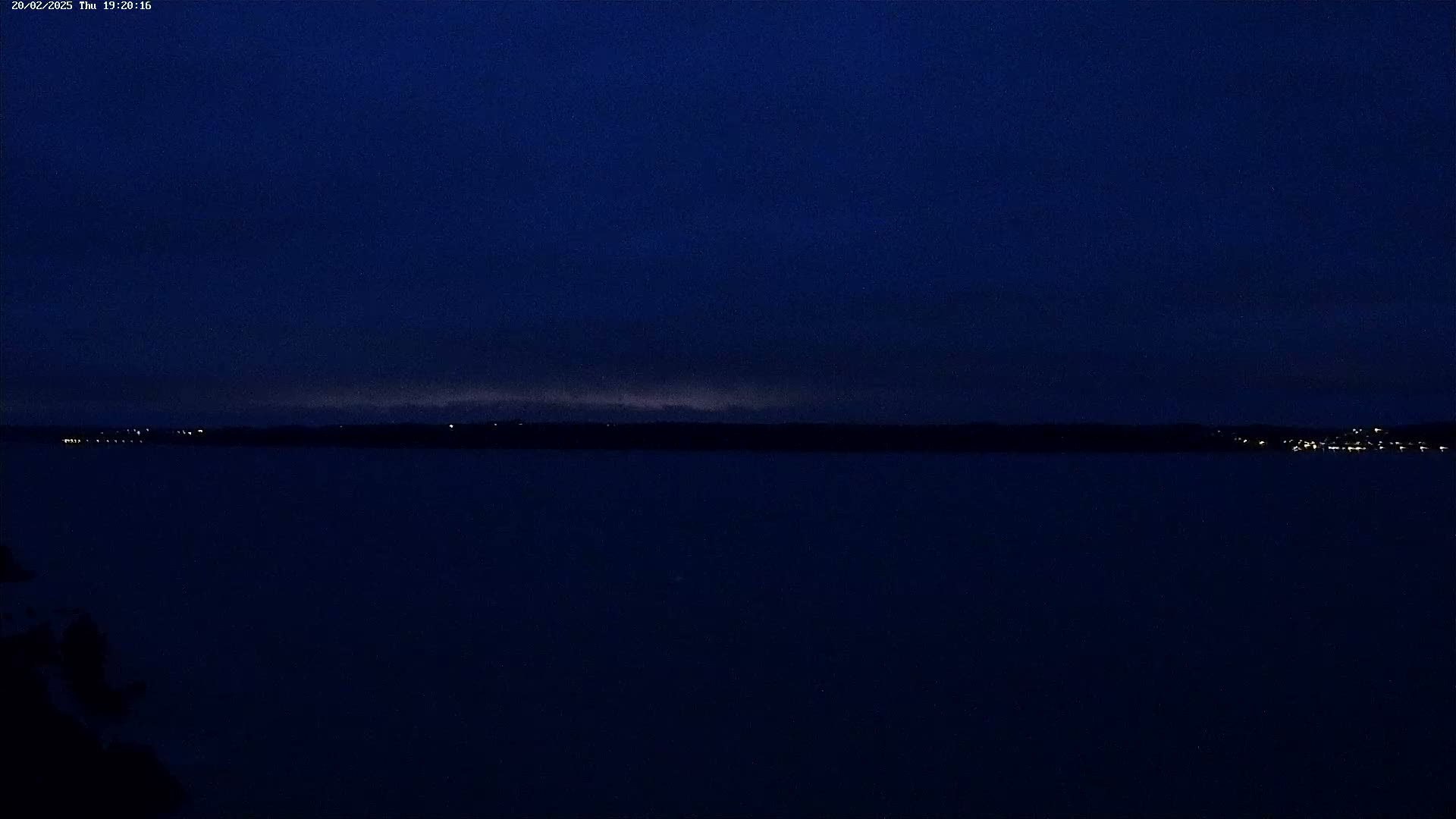 A dark, still body of water stretches to a distant shoreline faintly lit by scattered lights under a twilight blue sky.