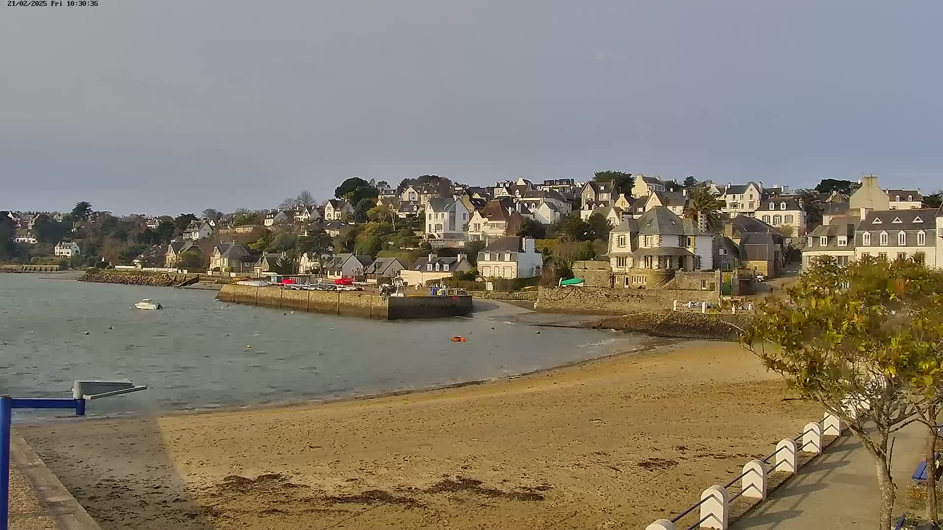 A sandy beach curves around a calm bay, with a small town of white buildings rising on a hillside in the background under a clear, light-grey sky.