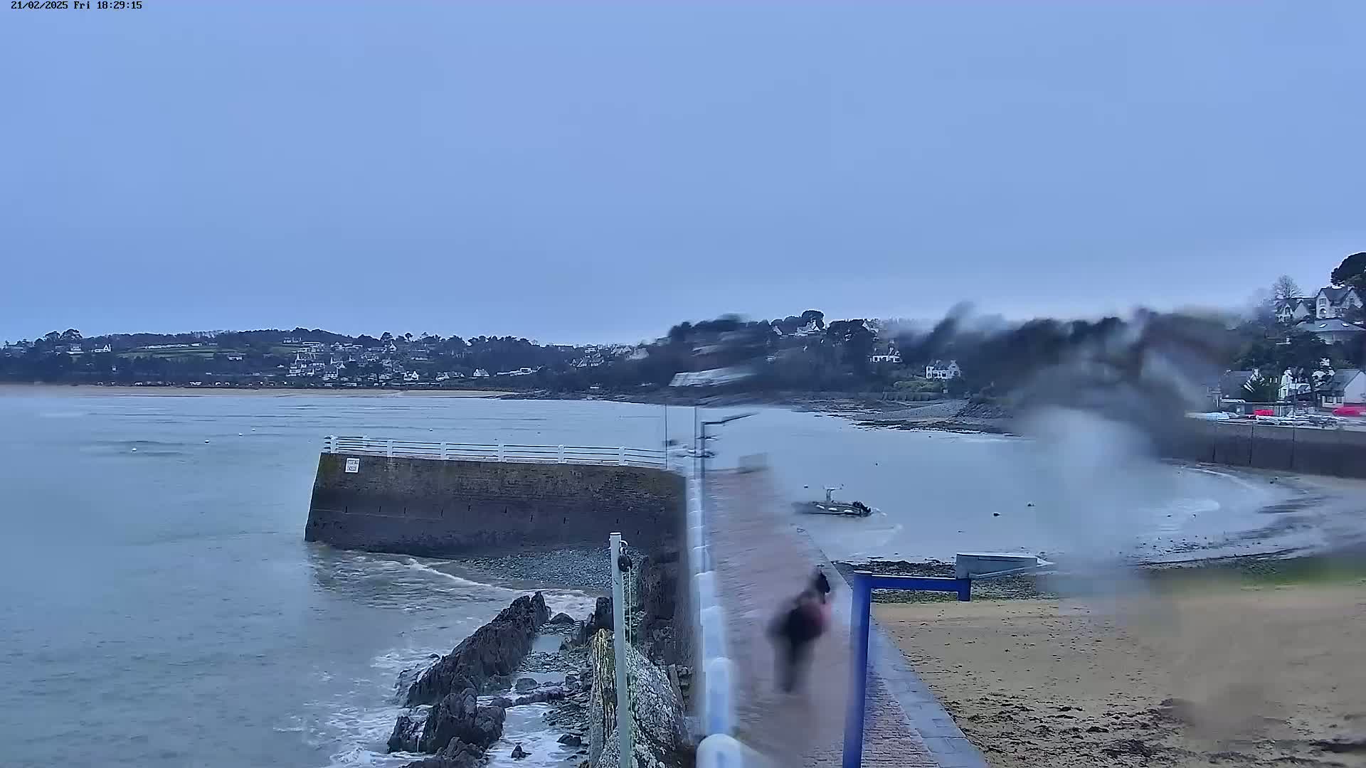 A person walks along a seawall overlooking a calm bay with a town in the distance under an overcast sky.