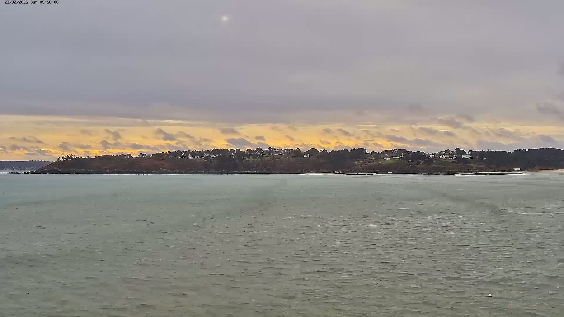 A calm body of water stretches out to a landmass with houses and trees under a cloudy sky with hints of sunset.