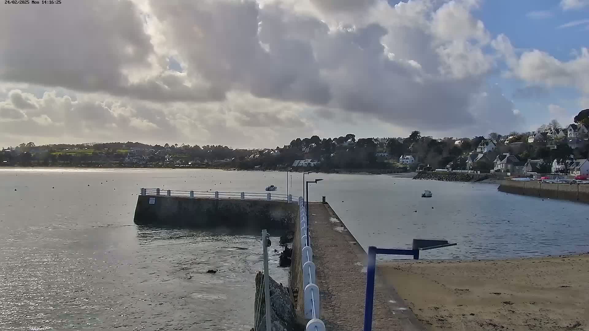 A partly cloudy day reveals a calm body of water with a small pier extending into it, leading to a sandy beach and a small town nestled on the opposite shore.