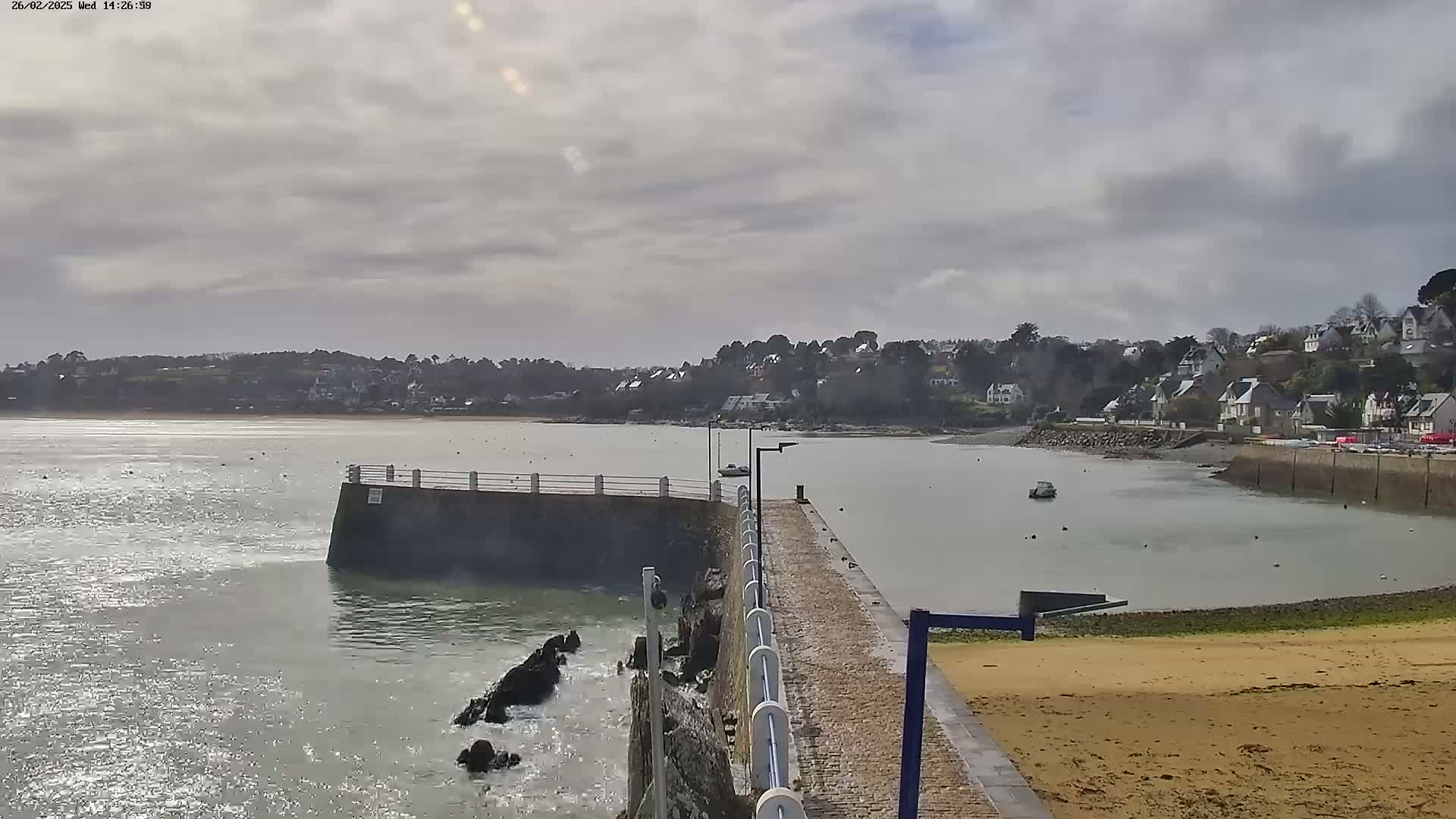 A stone pier extends into a calm body of water under a cloudy sky, with a small town visible on the opposite shore.