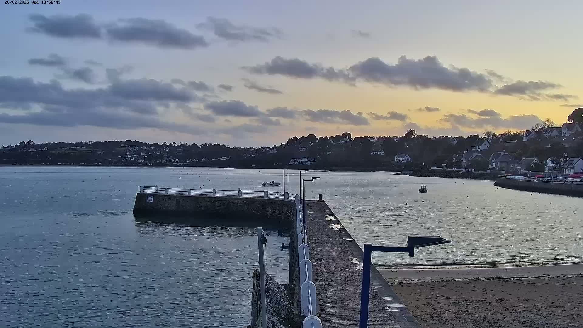 A stone pier extends into a calm body of water under a cloudy sunset sky, with a small town visible on the opposite shore.