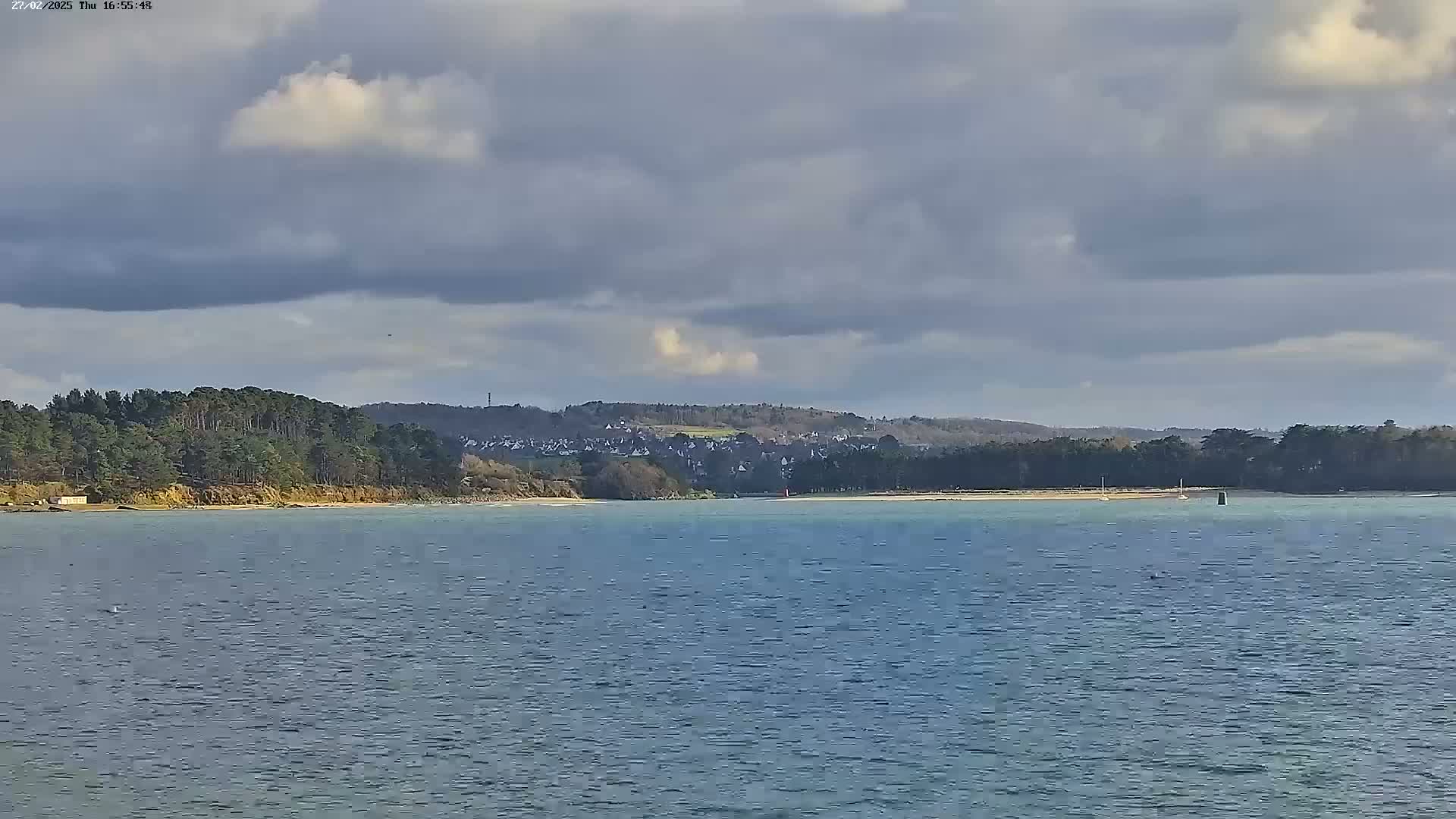 A body of calm water stretches towards a tree-lined shore, beyond which lies a village nestled against a hillside under a mostly cloudy sky.