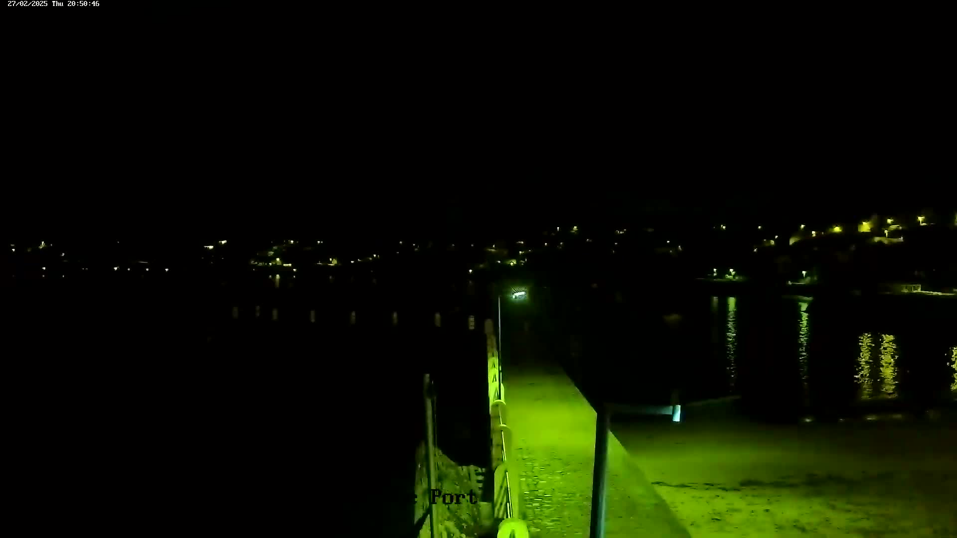 A nighttime view shows a lit walkway leading to a body of water reflecting the lights of a town across the water under a clear, dark sky.