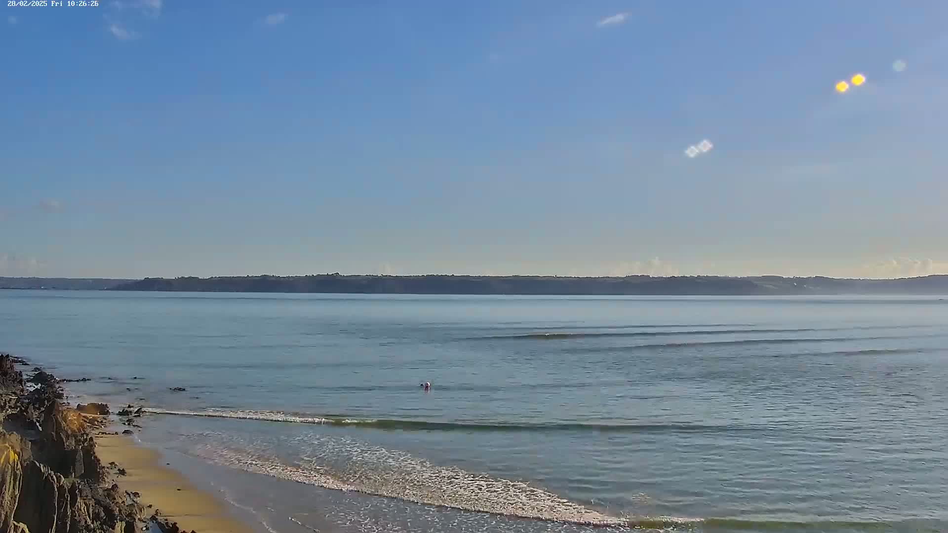 A sandy beach with small waves laps against rocky shores under a clear blue sky, with a distant landmass visible across calm water.