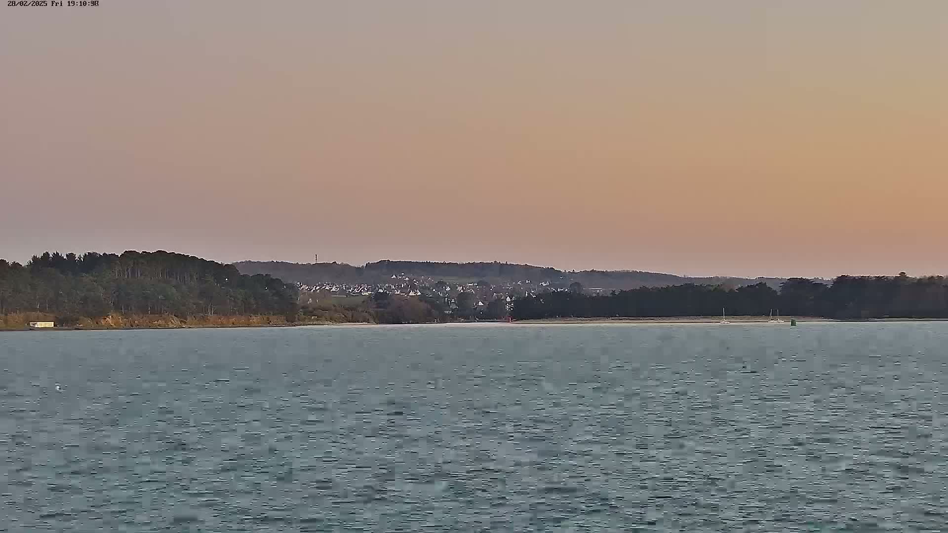 A calm body of water lies in front of a tree-lined shore, beyond which sits a small town under a pale pink sunset sky.
