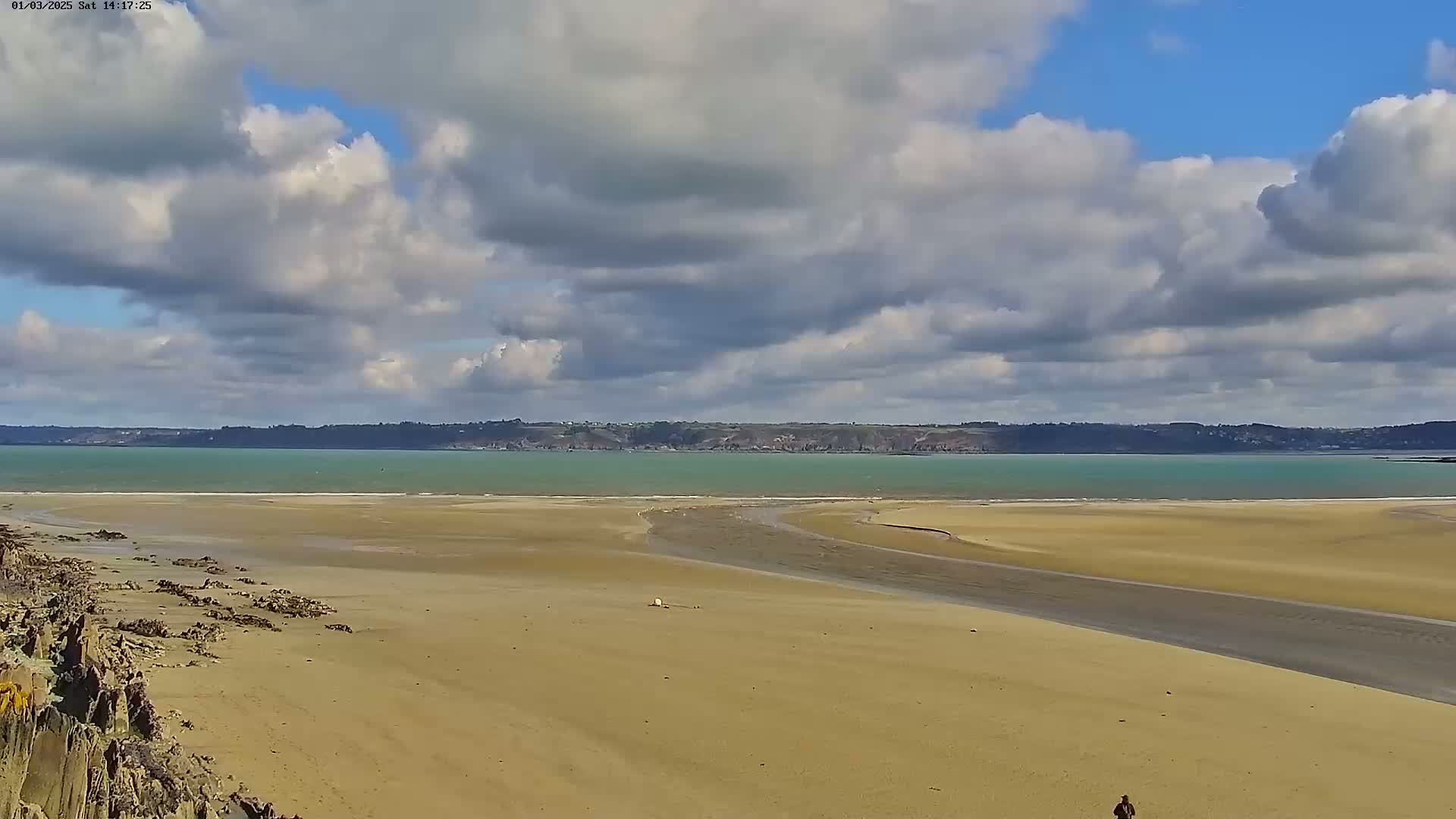 A wide sandy beach stretches toward a calm, teal-colored bay under a partly cloudy sky.