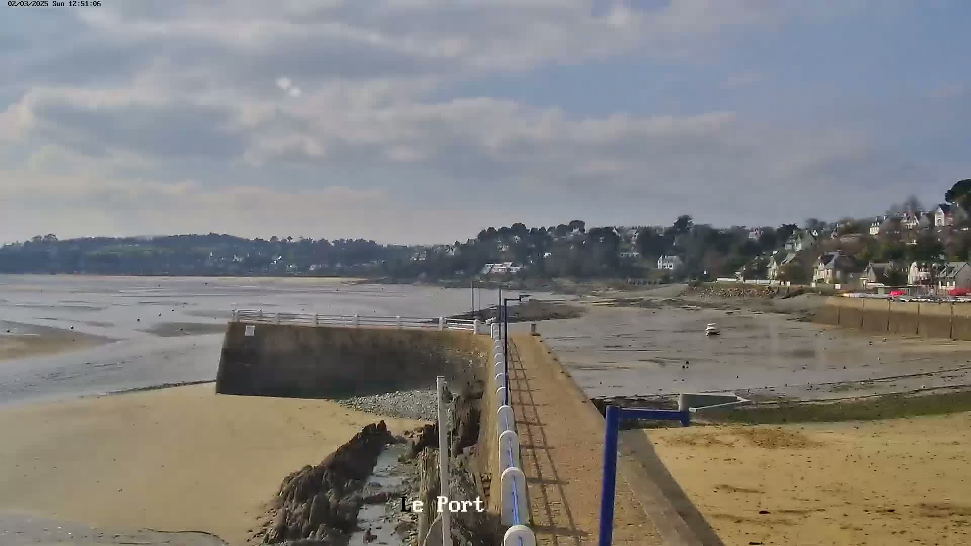 A partly cloudy day reveals a sandy shoreline with a low tide exposing a wide expanse of mudflats, a small harbor, and a distant village nestled among trees.