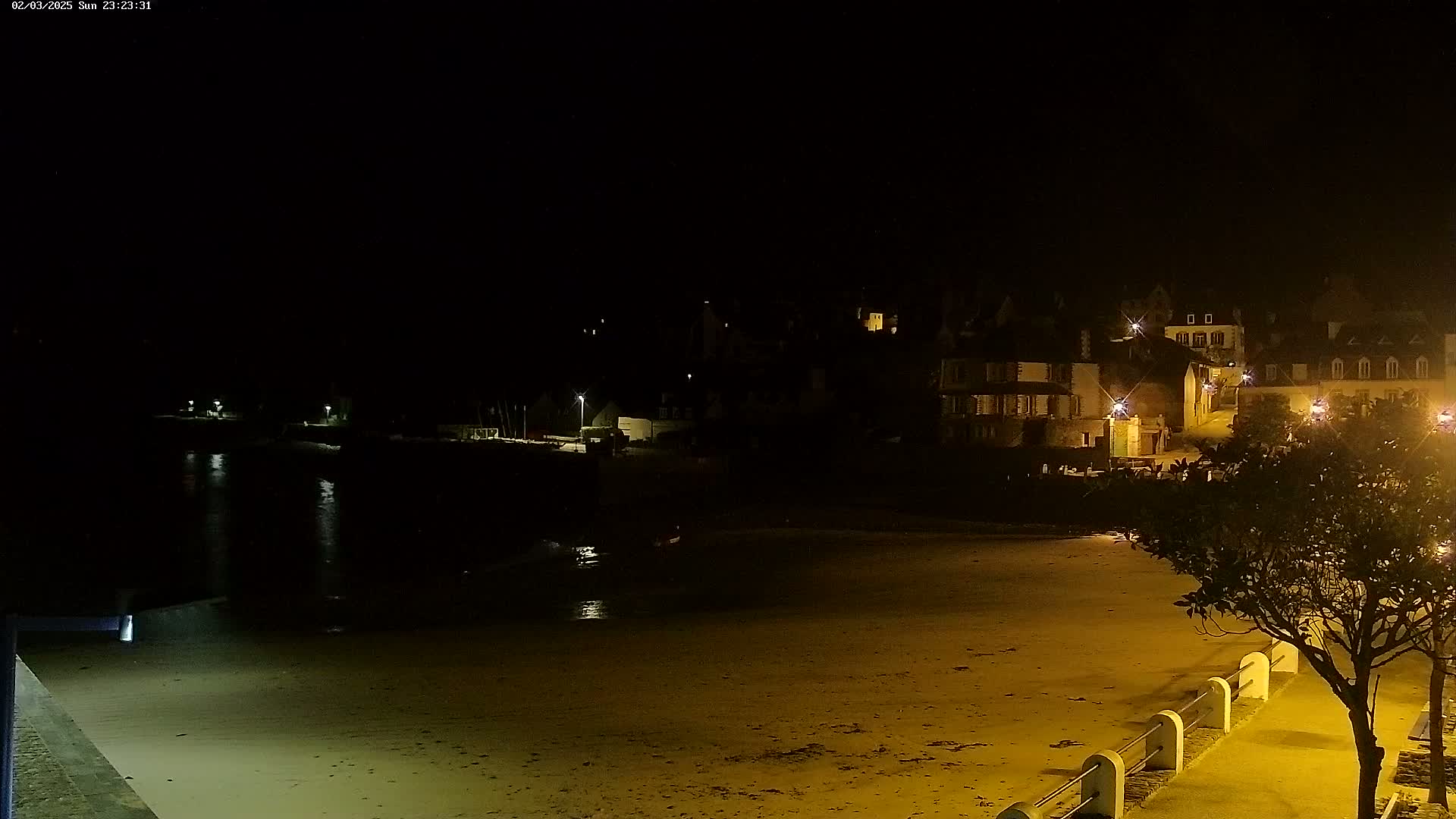 A dark, nighttime beach scene shows a sandy shoreline, calm water reflecting lights, and a village of buildings illuminated with streetlights in the background.