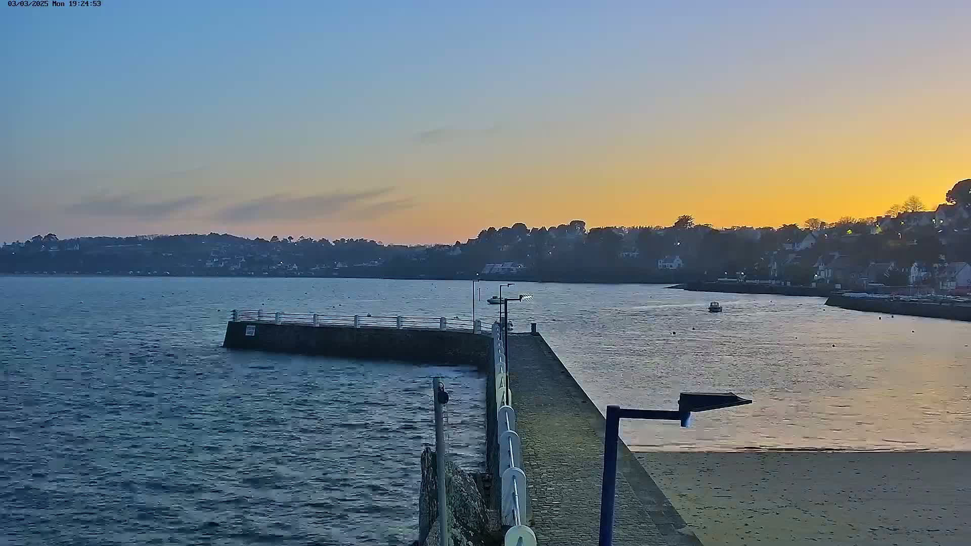 A small pier extends into a calm body of water at sunset, with a town visible in the distance under a clear sky.