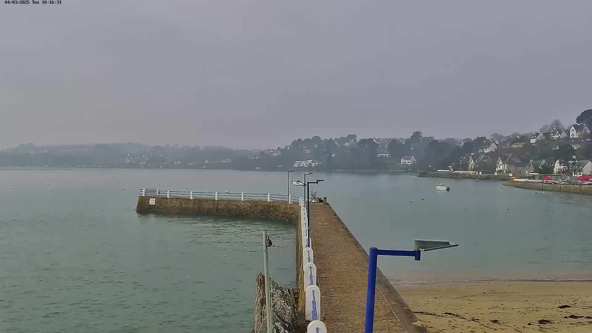 A hazy, overcast day reveals a small stone pier extending into calm water, where a few boats are visible beyond a shoreline of houses and trees.