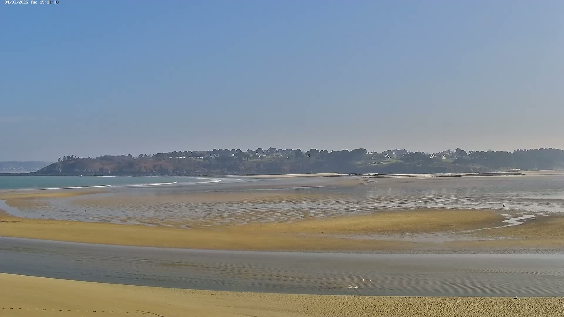 A sandy beach stretches out to a calm bay, where a distant shoreline with a line of trees and buildings is visible under a clear blue sky.