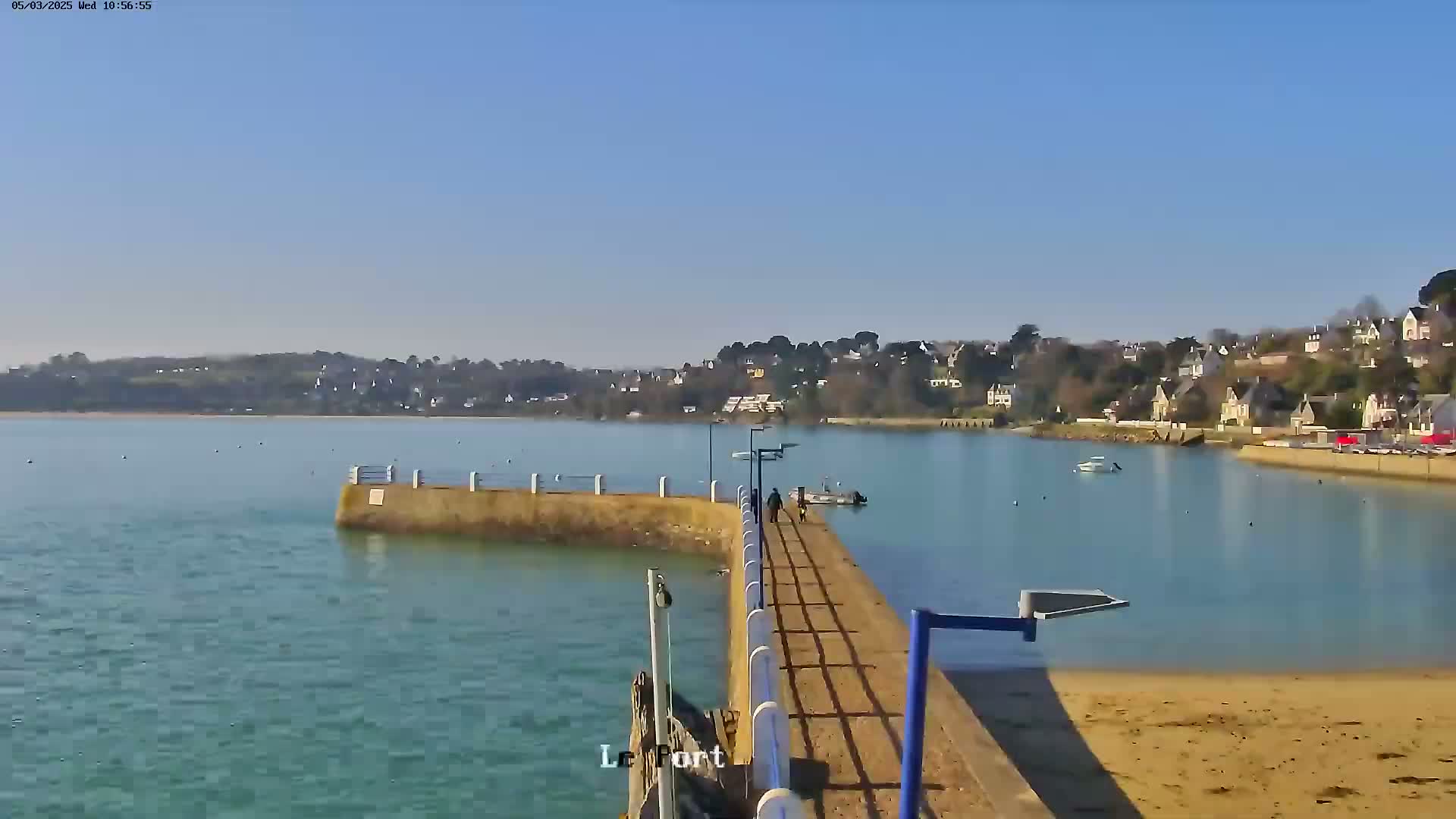 A long pier extends into a calm, light-blue body of water on a sunny day, with a small town visible across the water.