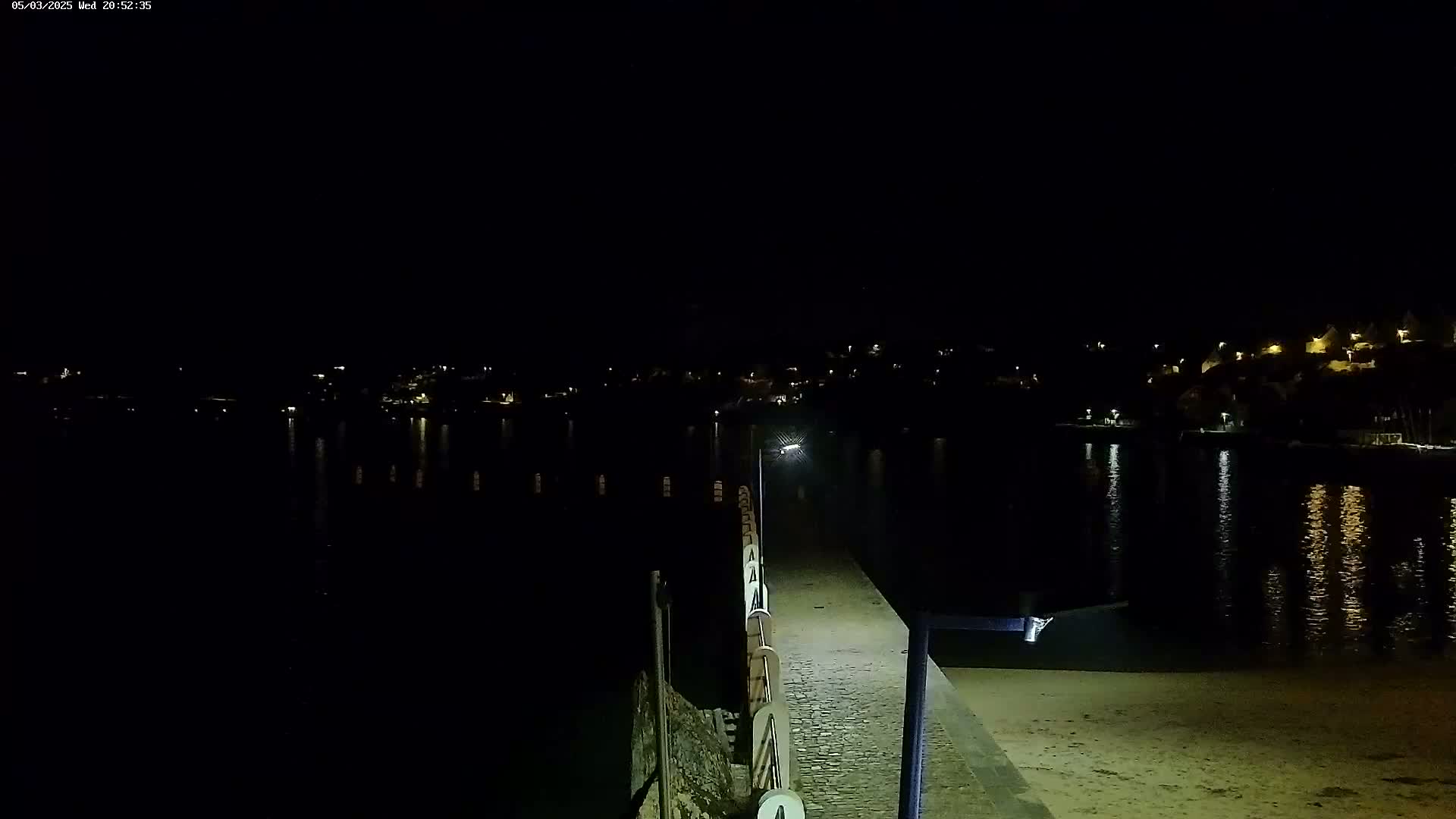 A nighttime view of a paved walkway along a body of water, with dimly lit buildings visible across the water.