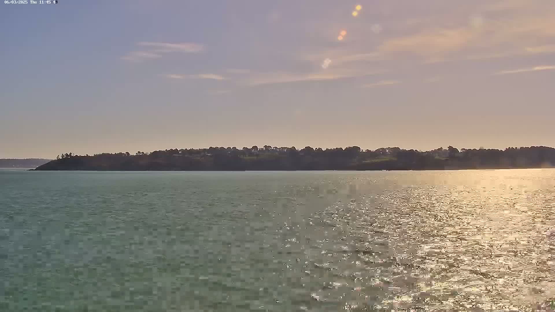 A tranquil body of water reflects the sunlight under a mostly clear sky, with a tree-lined shoreline in the distance.