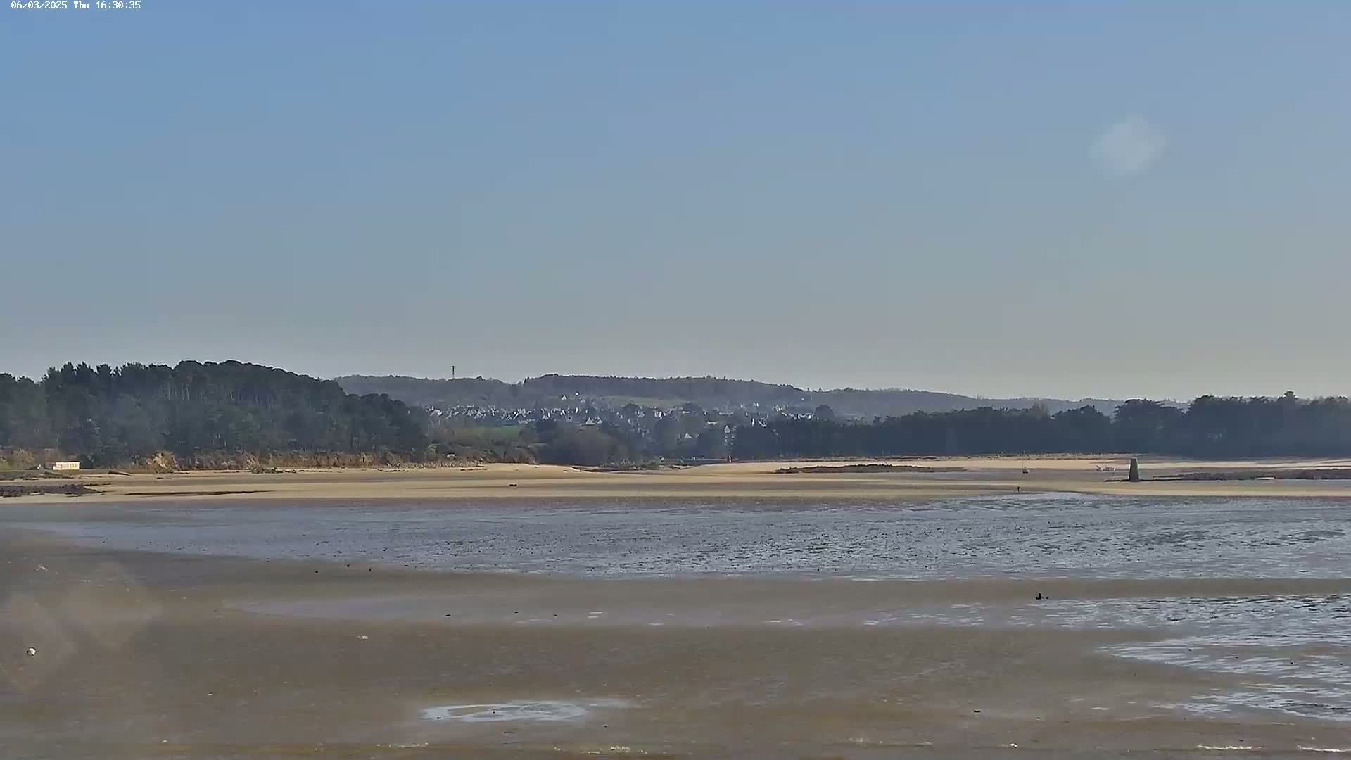 A wide, sandy beach stretches out to calm, shallow water under a clear, light blue sky, with a line of trees and a distant village visible in the background.