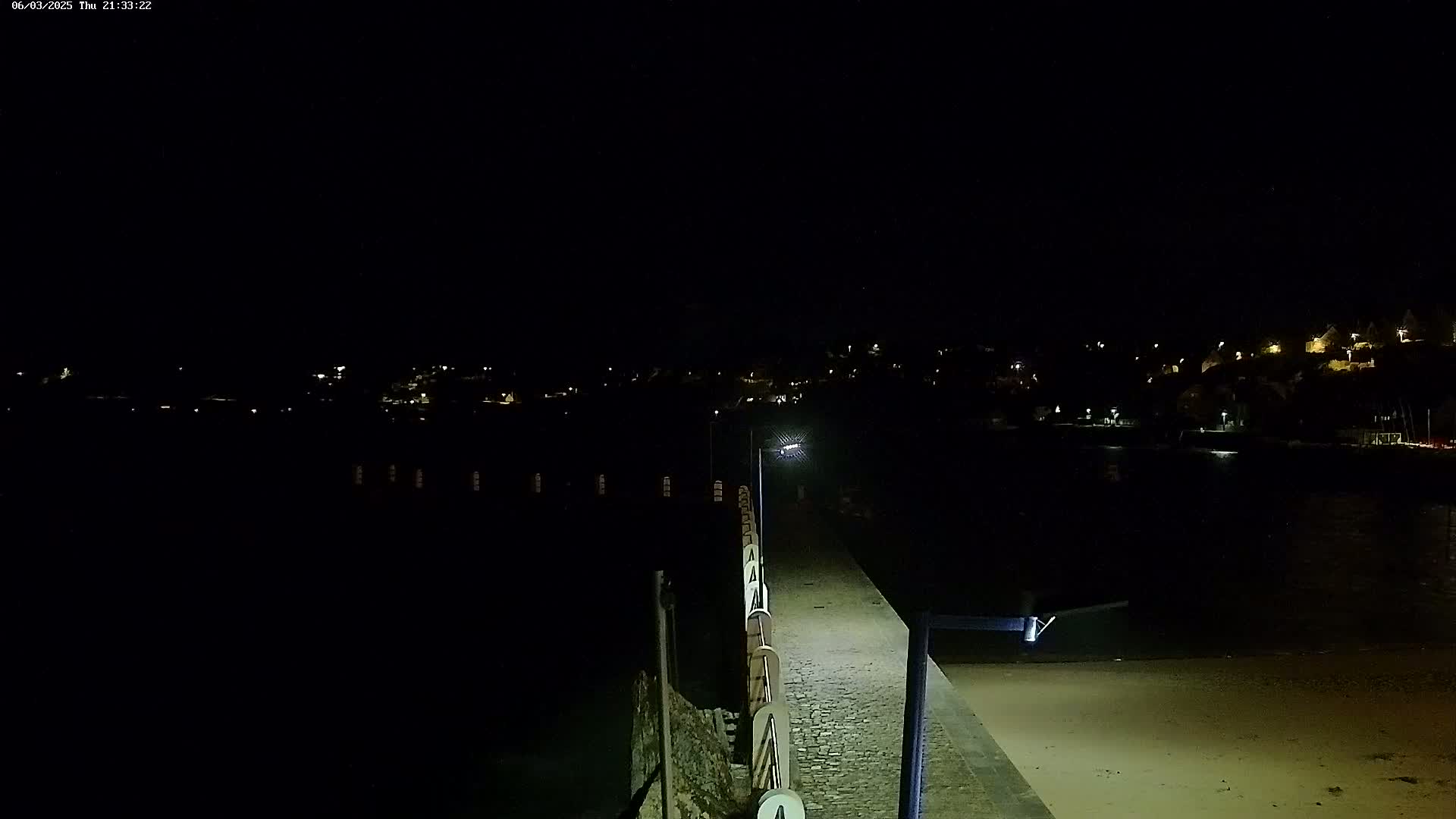 A nighttime view shows a paved walkway extending from a beach toward a dimly lit town across a body of water.