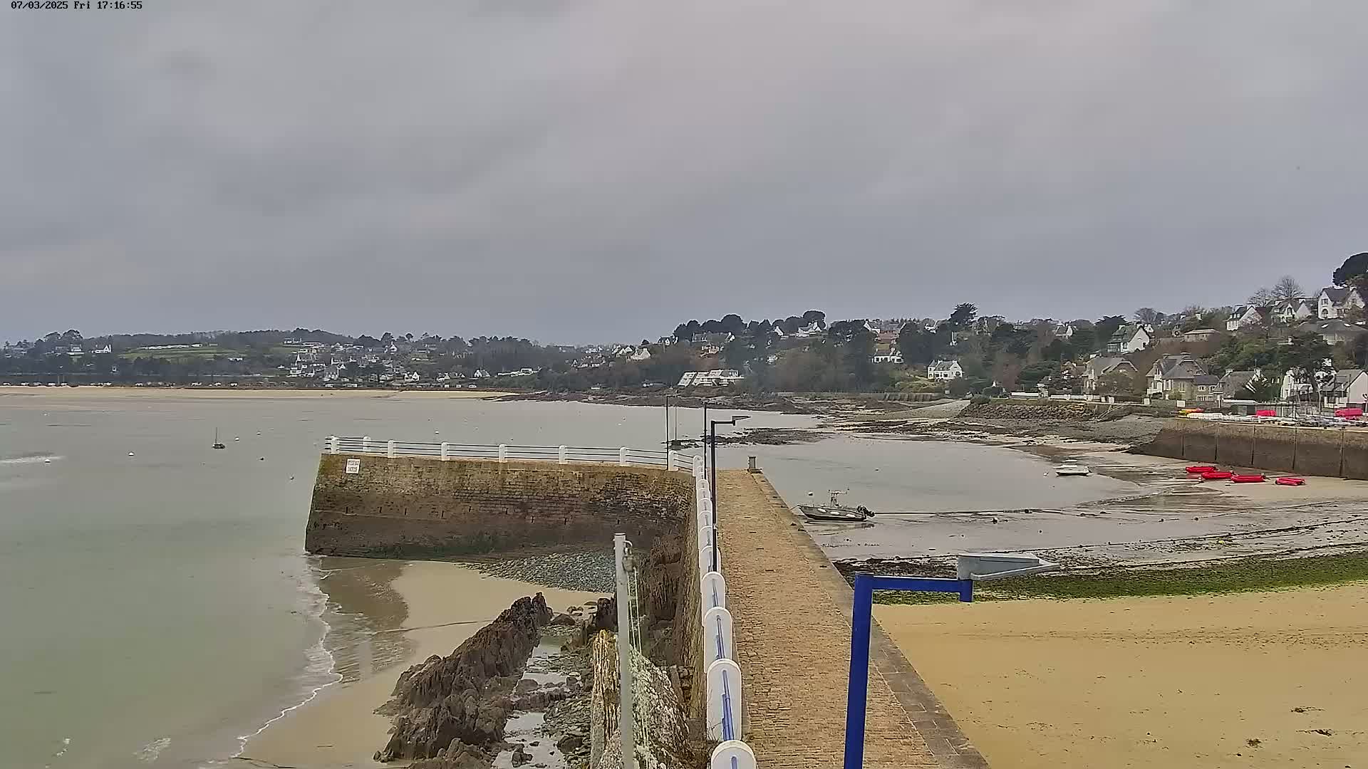 A stone pier extends into a calm, gray-skied bay next to a sandy beach, with a small town visible across the water.