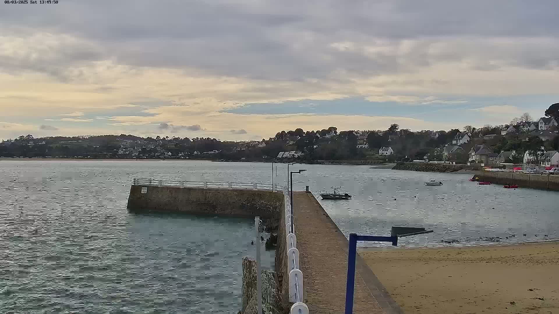 A small pier extends into a calm, gray body of water, with a small town and houses visible on the other side under a partly cloudy sky.