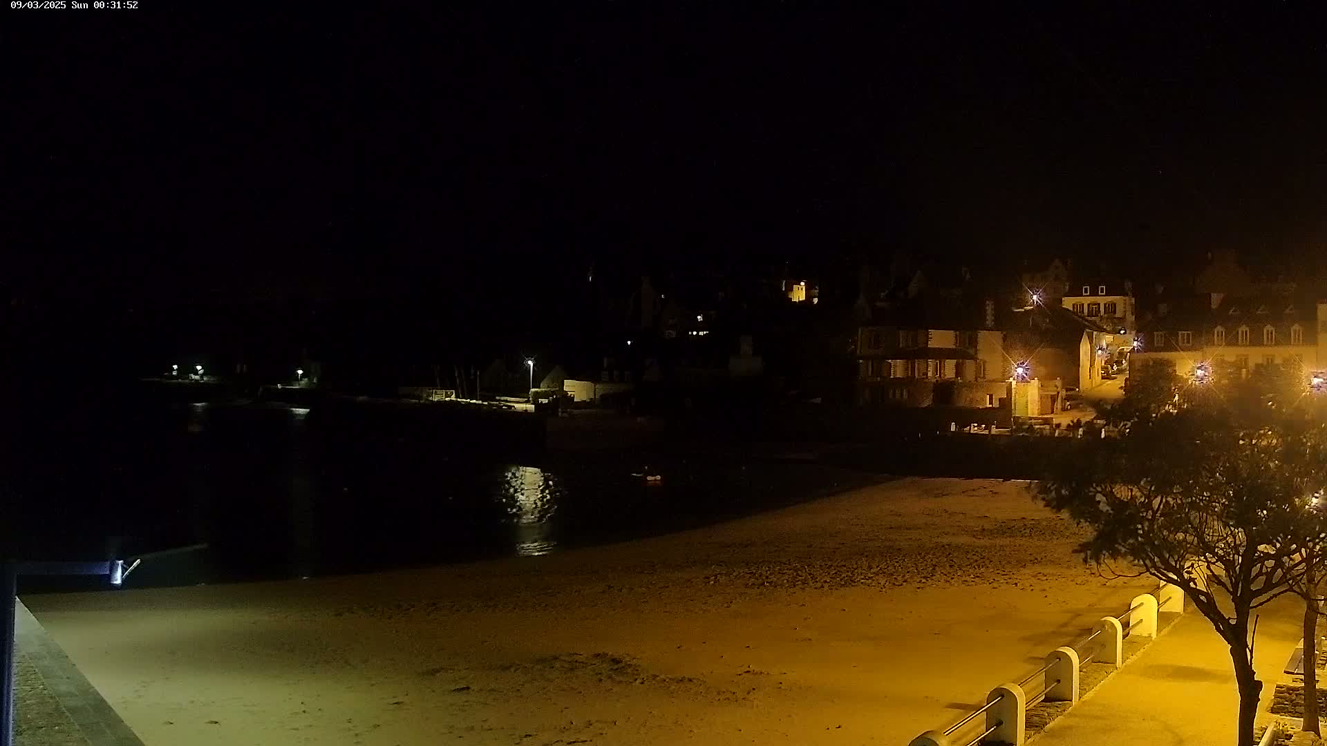 A nighttime view of a sandy beach and calm water, with a village of lit buildings across the water in the background.