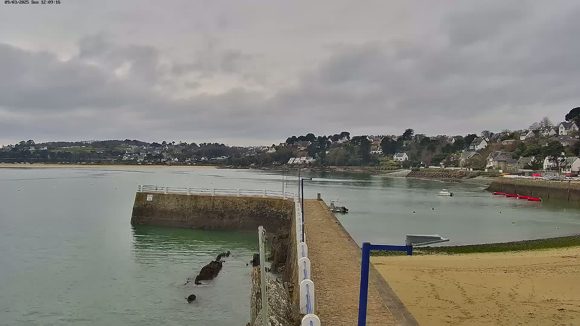 A stone pier extends into a calm body of water under an overcast sky, overlooking a shoreline dotted with houses and trees.