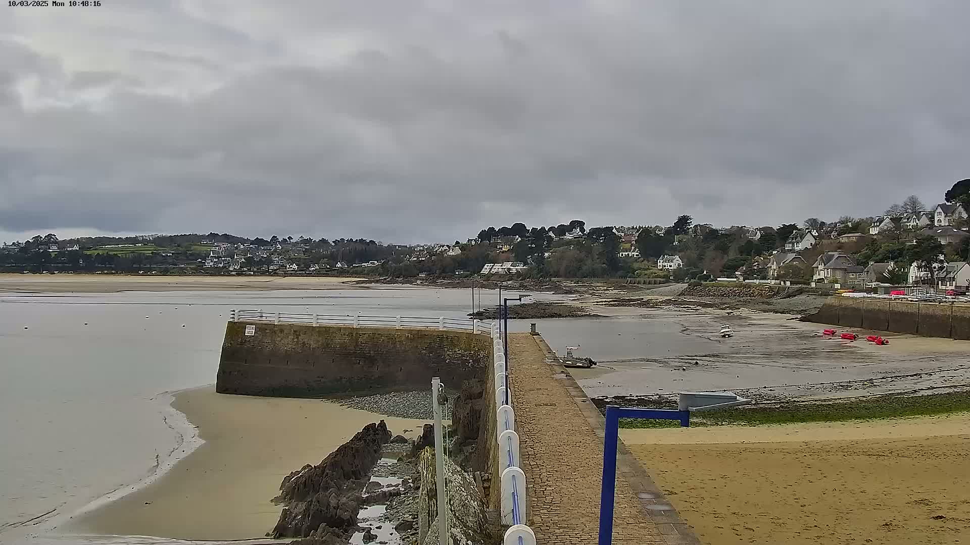 A low-tide view of a sandy beach and calm bay, under an overcast sky, shows a small town on the opposite shore with a stone pier extending into the water.