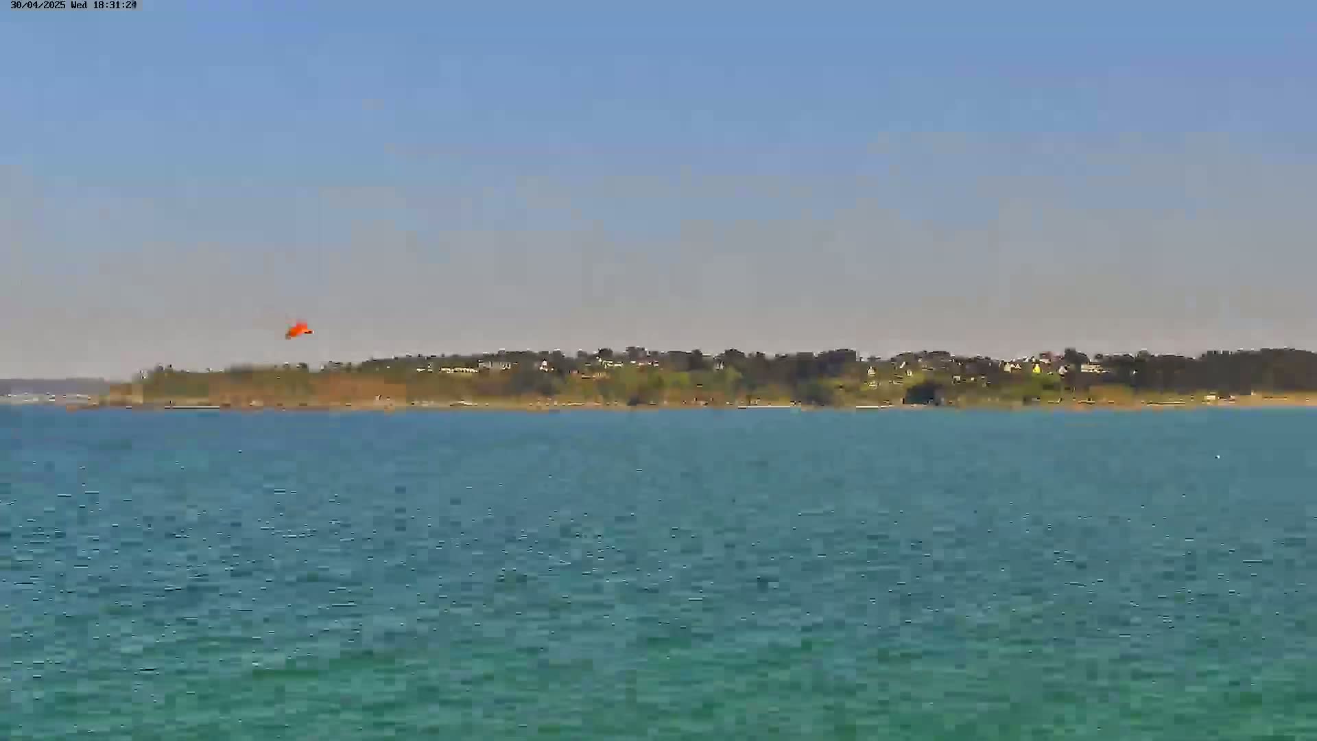 A small orange object is visible in the sky above a calm body of teal water that stretches to a tree-lined shore on a clear day.