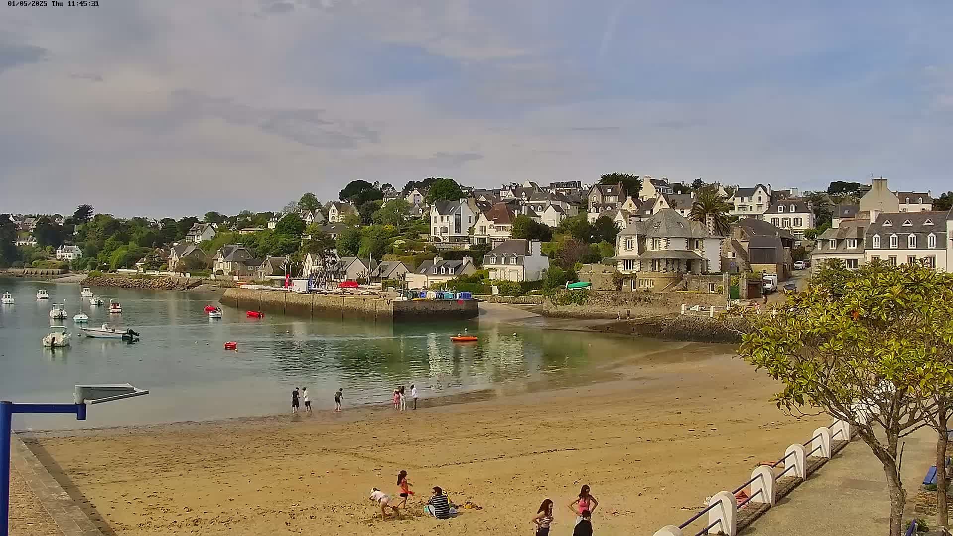 A sandy beach with people relaxing and playing, boats in the calm water of a harbor, and a picturesque village of white houses nestled on a hillside under a partly cloudy sky.