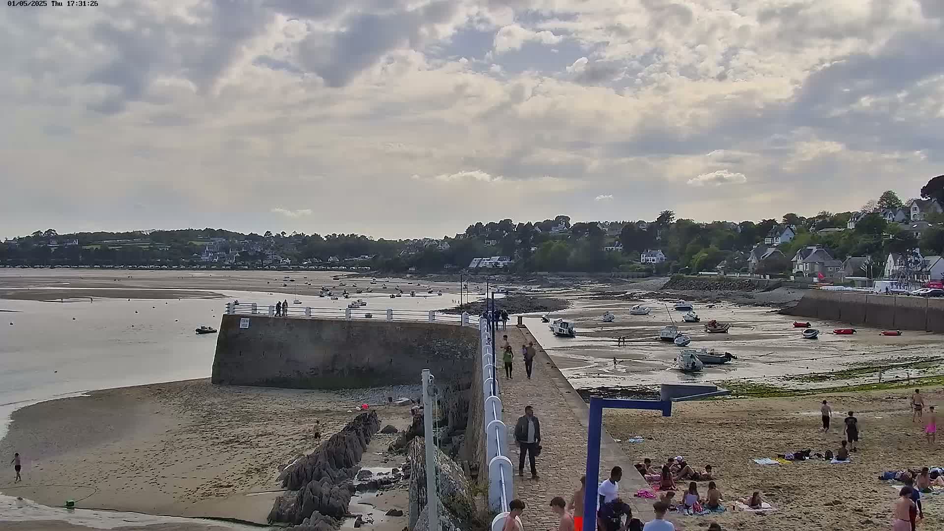 A sandy beach with people sunbathing and walking on a pier, boats resting on exposed mudflats at low tide under a partly cloudy sky.