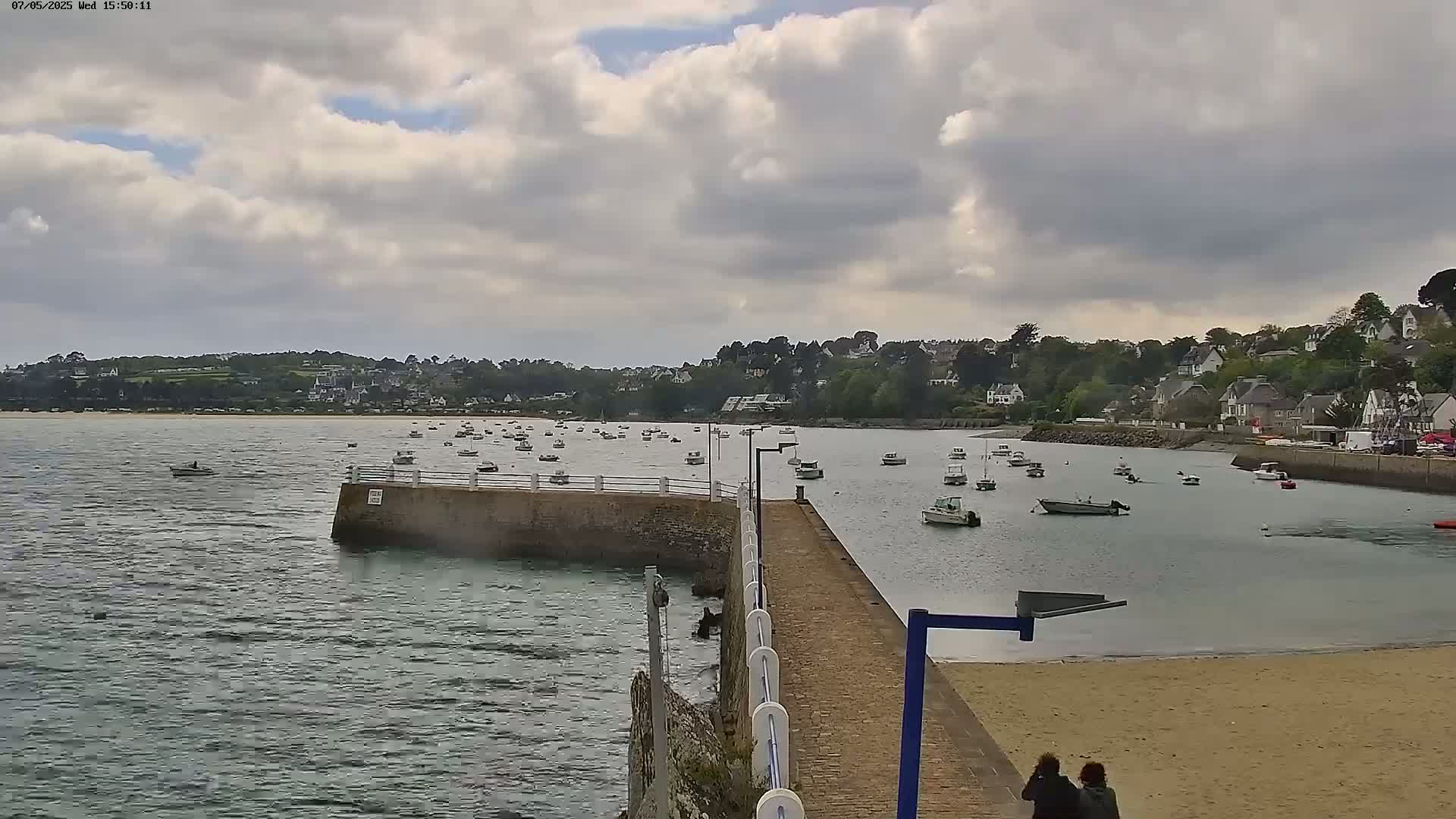 A paved jetty extends into a calm bay filled with numerous small boats under a partly cloudy sky, with a sandy beach and a small town visible in the background.