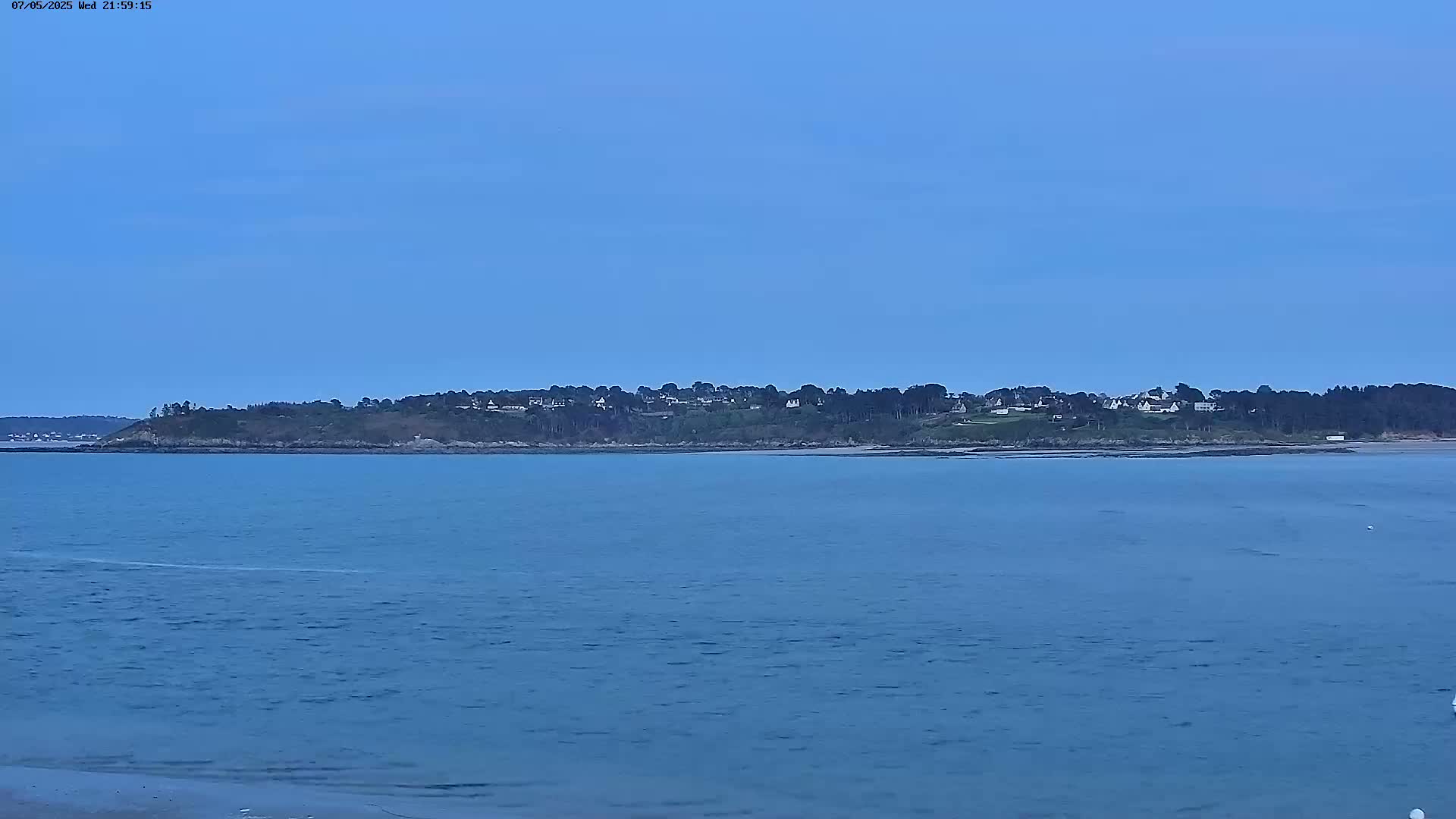 A tranquil, blue body of water stretches towards a distant shoreline where a small town is nestled amidst a line of dark green trees under a clear, twilight sky.
