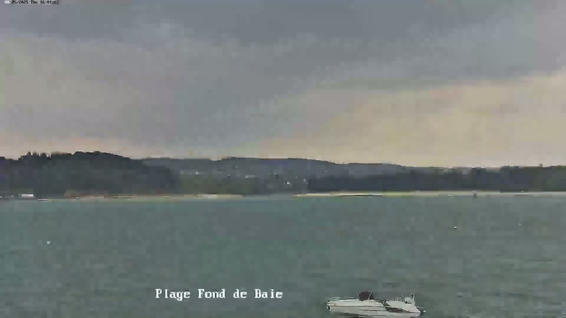A small motorboat sits in calm, grey water in front of a tree-lined shore under an overcast sky.
