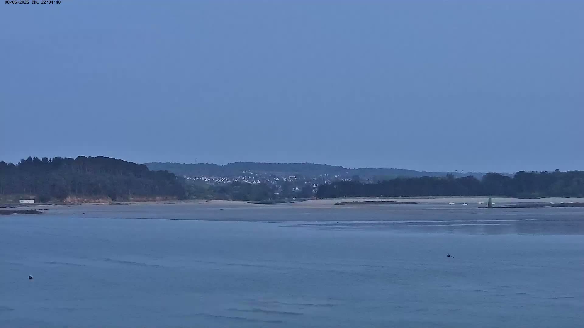 A calm body of water at dusk shows a sandy shore with a line of trees and a small town in the background.