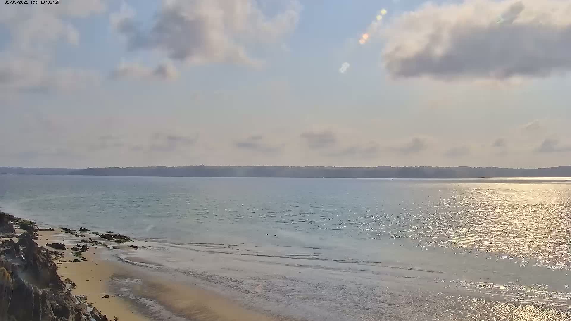 A partly cloudy sky overlooks a calm, sunlit body of water with a sandy shoreline and rocky outcroppings.