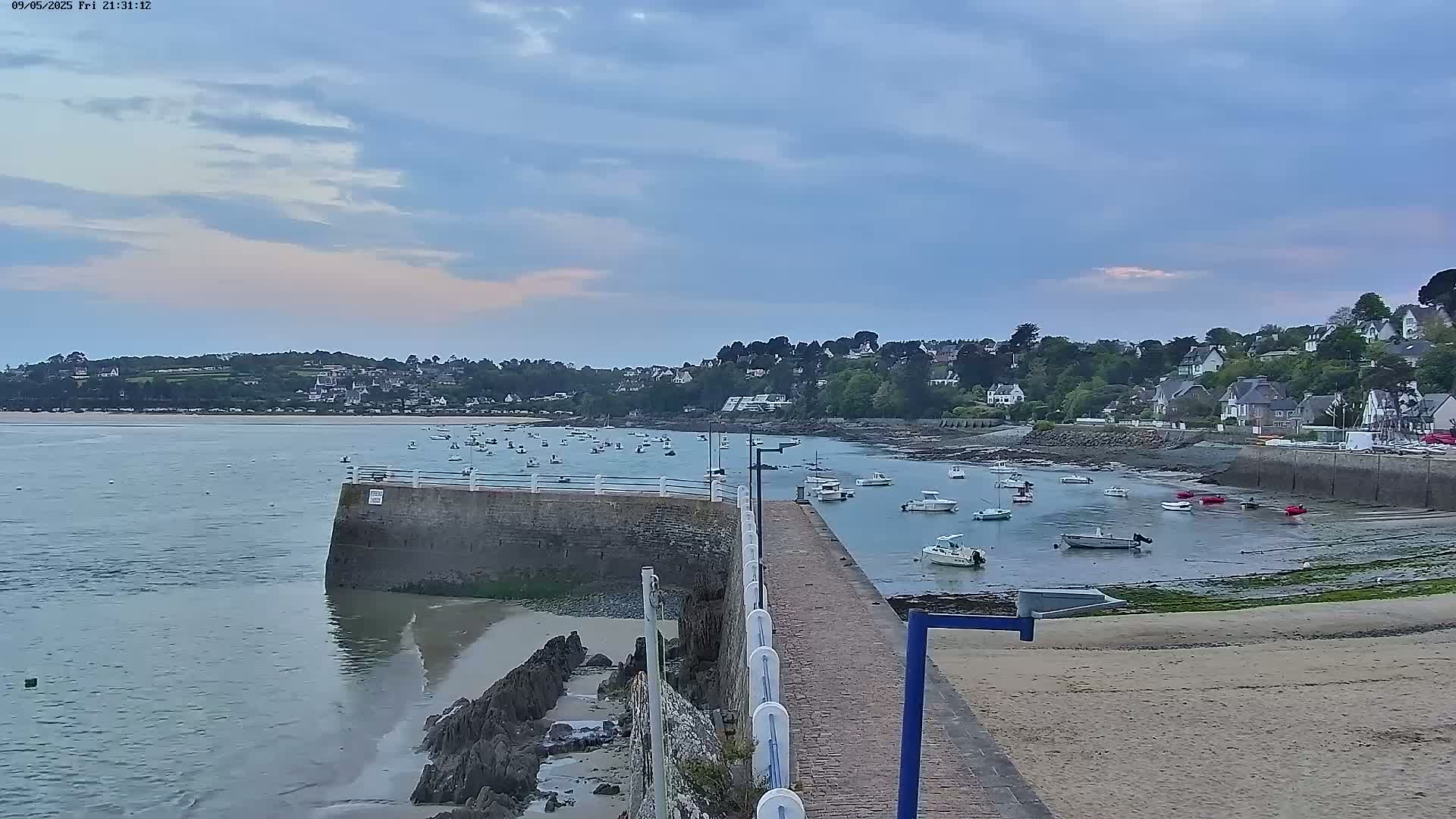 A calm, overcast evening scene shows a stone pier extending into a harbor filled with small boats,  with a residential area on a hillside in the background.