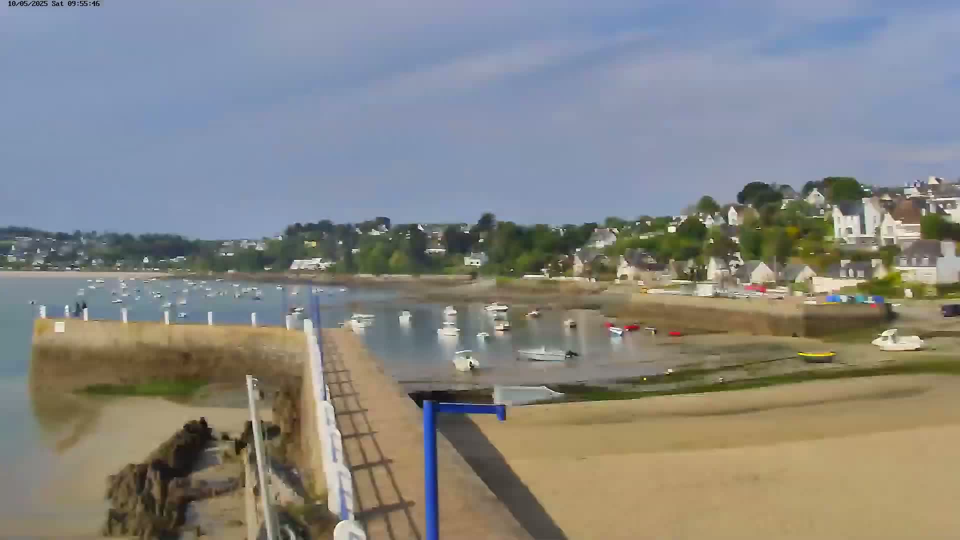 A sunny day reveals a harbor filled with numerous boats at low tide, nestled beside a small town with houses lining a gently sloping hillside.