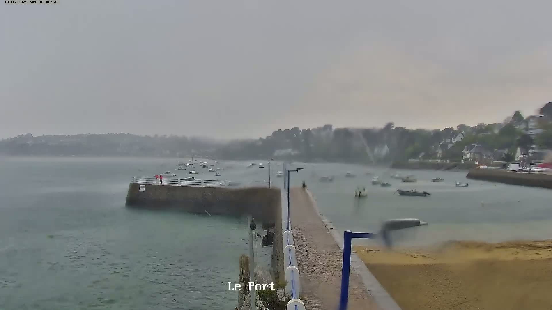 A harbor filled with numerous small boats is viewed from a stone pier on a heavily overcast day with wind and apparent rain.