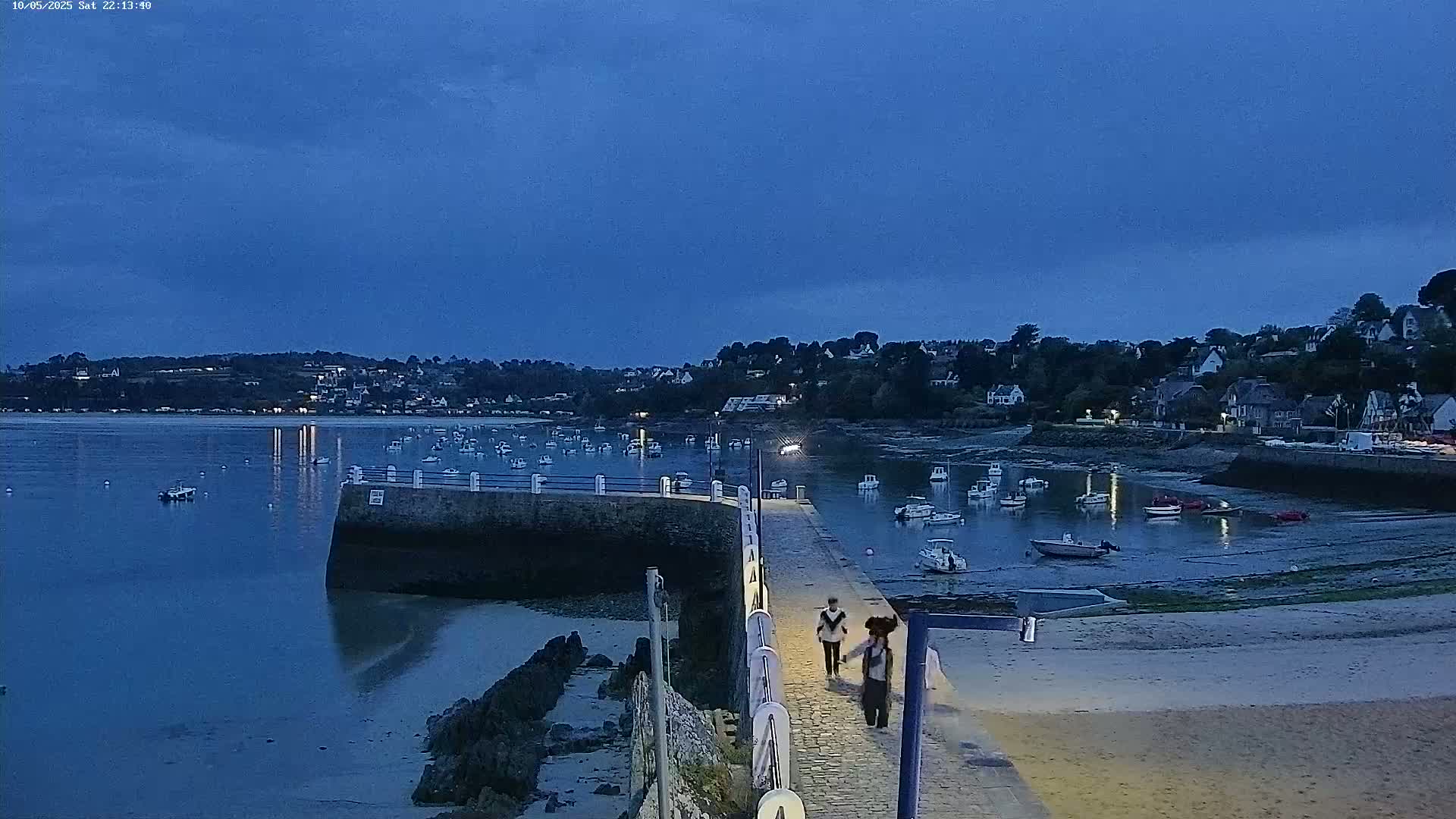 Two people walk down a pier at dusk toward a small harbor filled with boats, under a clear blue-grey sky.