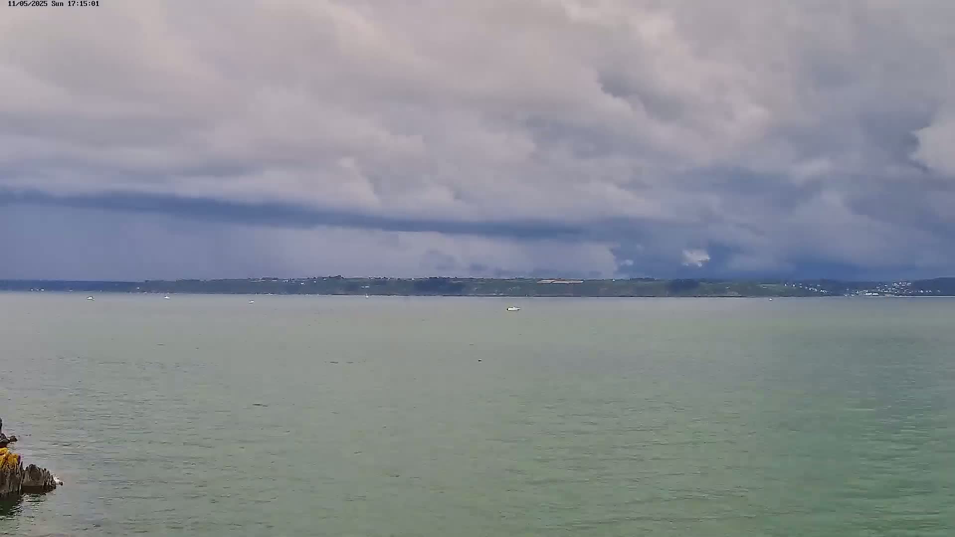 A body of calm water stretches to a distant shoreline under a mostly cloudy sky with dark, heavy clouds looming overhead.