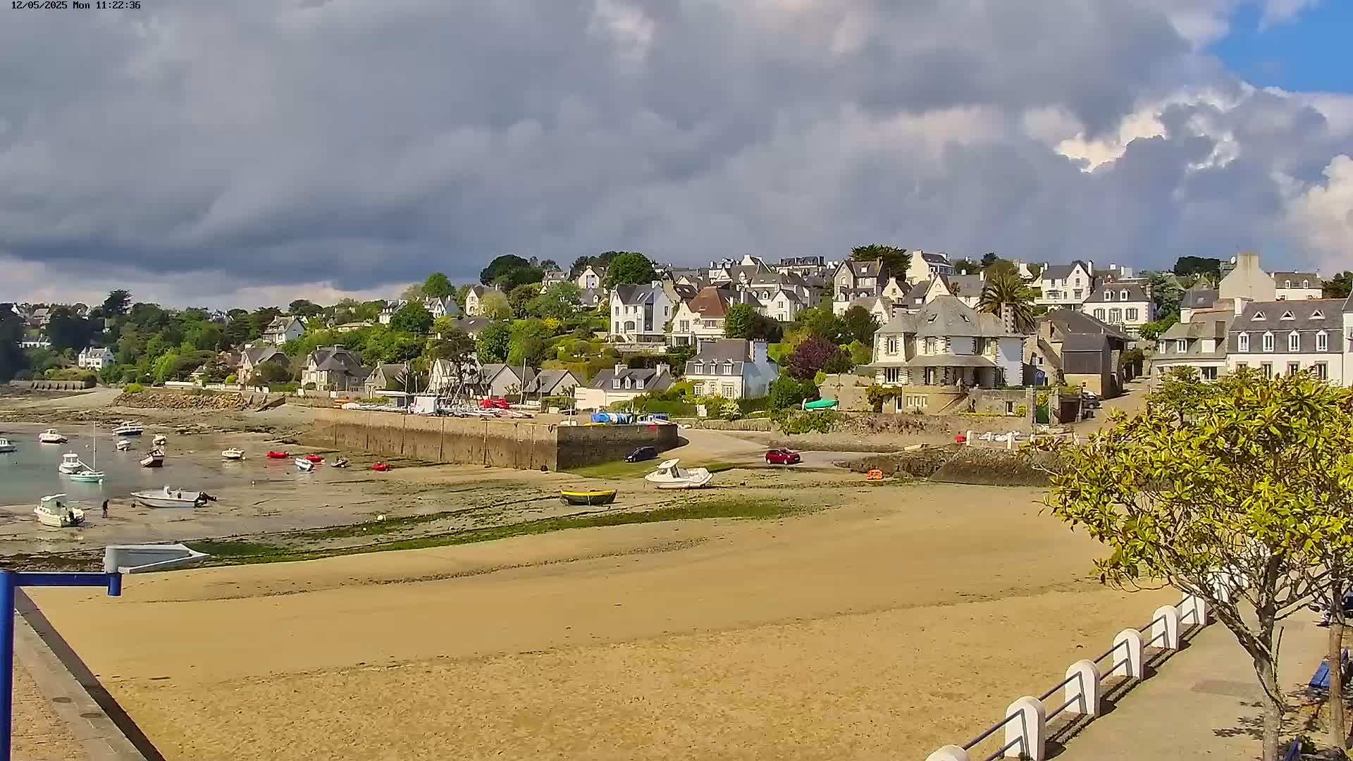 A sandy beach with several small boats rests at low tide in front of a hillside village under a partly cloudy sky.