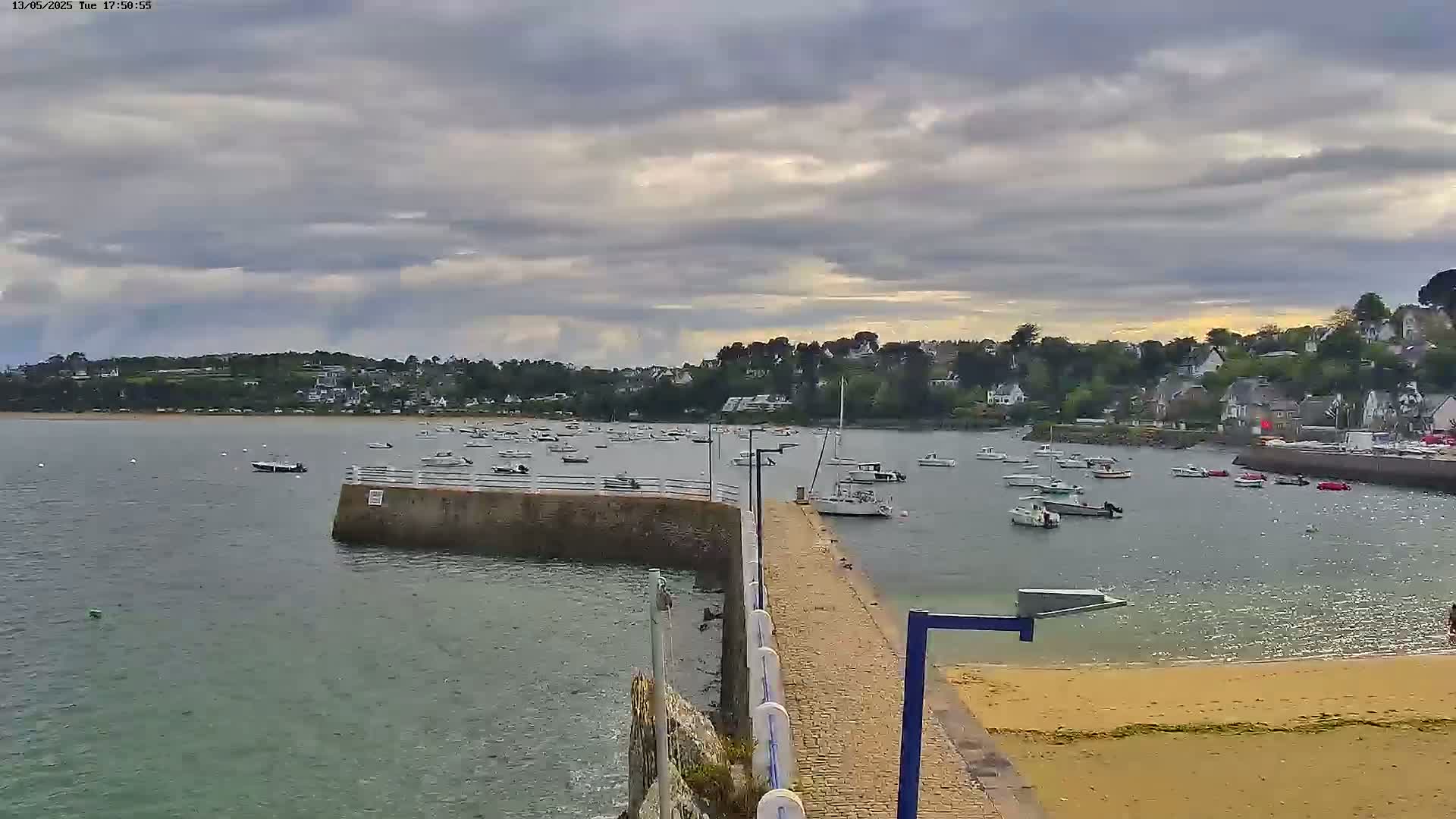 A calm bay with numerous boats is seen from a stone jetty leading to a sandy beach under a cloudy sky.